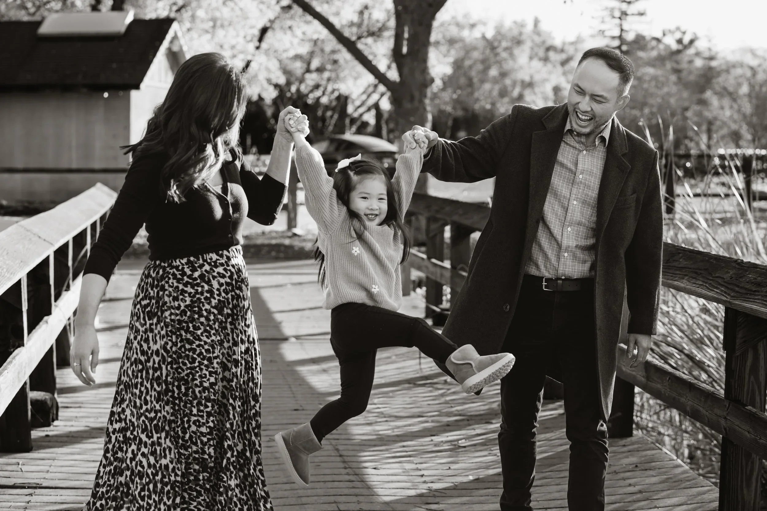 In black and white, a mother and father each hold one of their daughter's hands and swing her mid-step, one leg kicked up, on a wooden bridge.