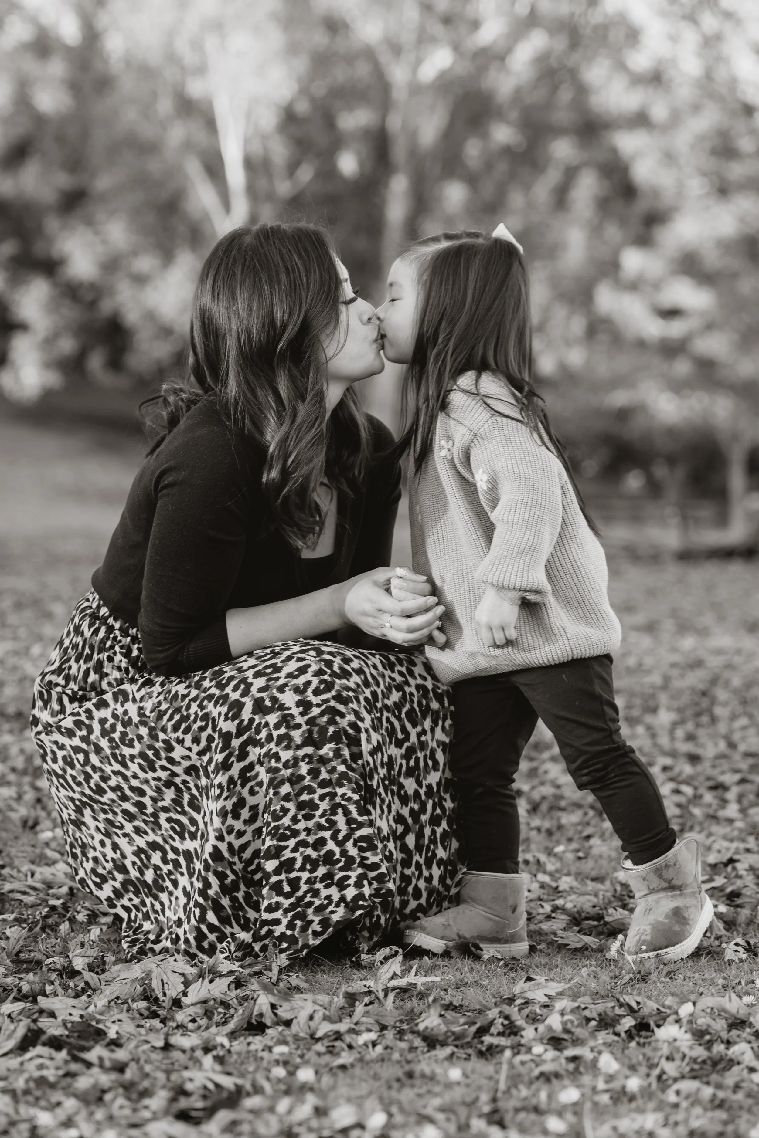 In black and white, a mother kneels to kiss her daughter on the lips while holding both her hands, surrounded by fallen leaves.