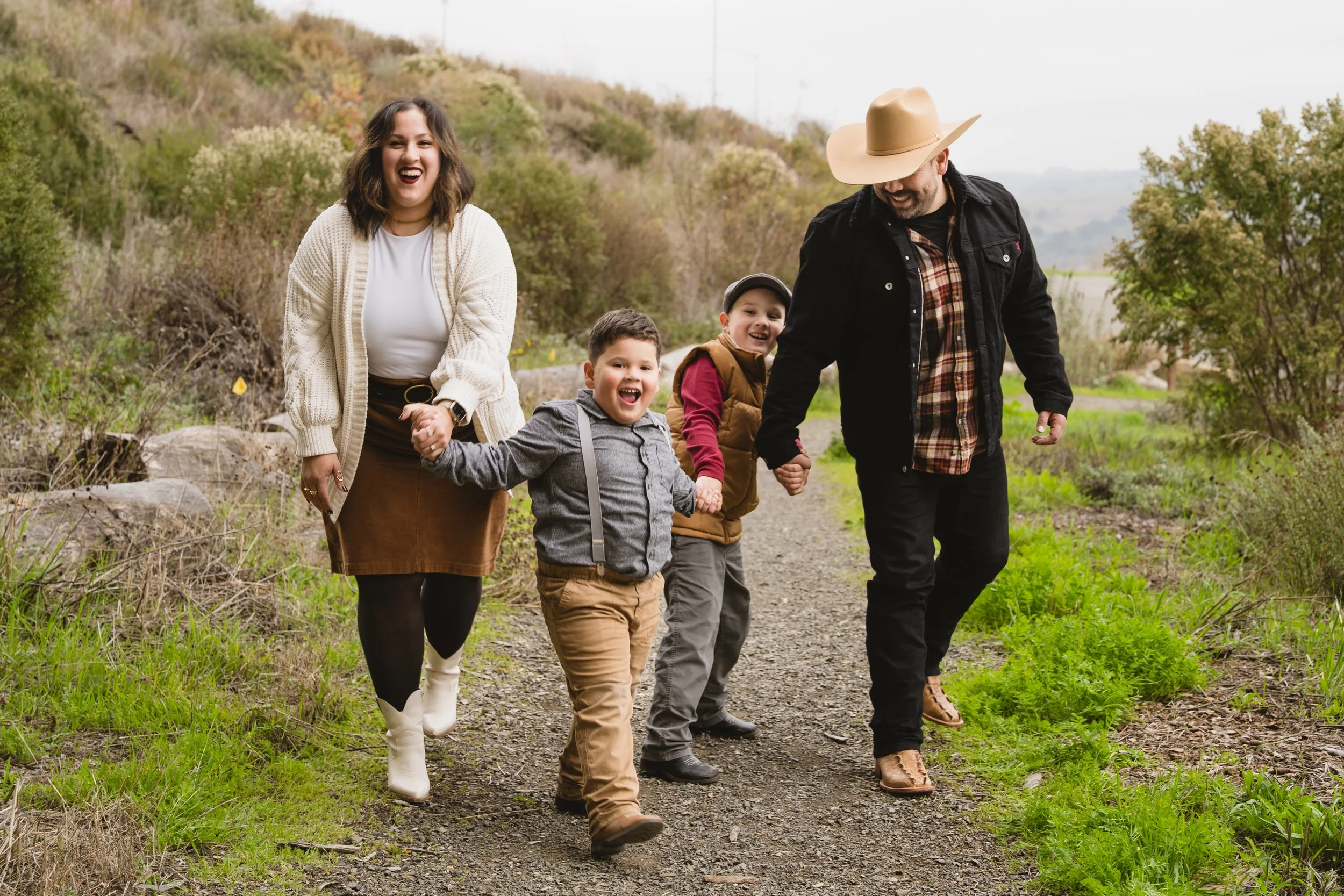 Group of four people, two adults and two children, walking hand-in-hand along a dirt trail surrounded by green foliage and hills, smiling and enjoying a day outdoors.