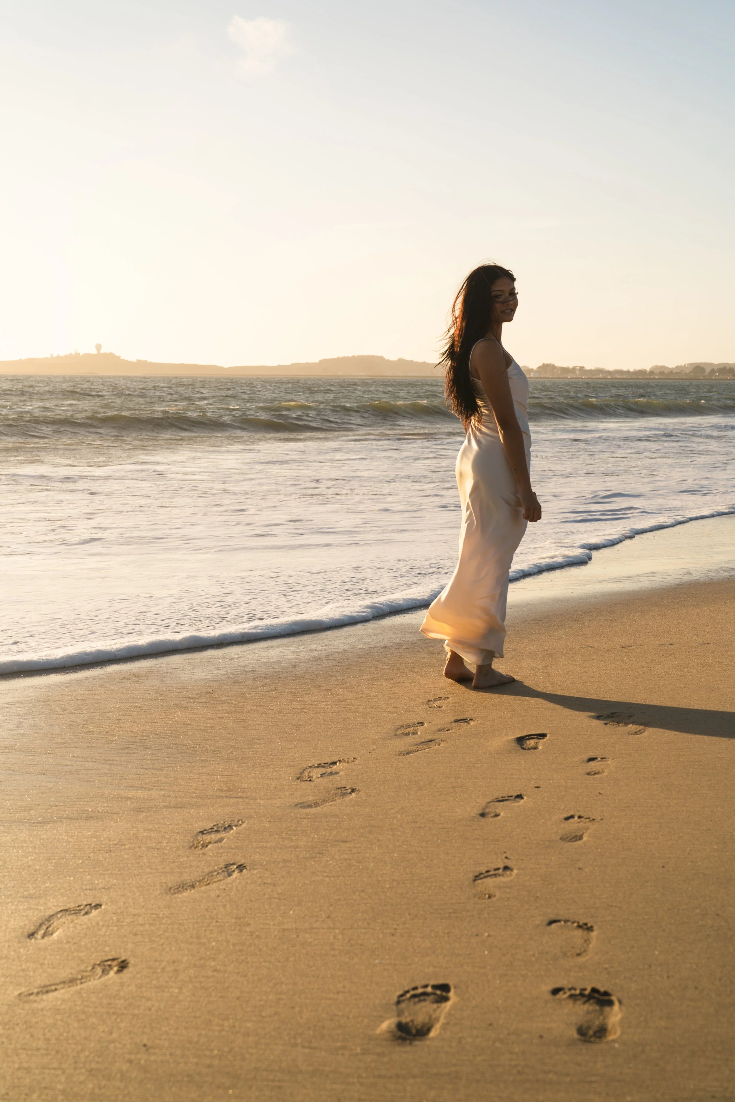 Senior walking toward the ocean at sunset, her footprints trailing across the sand in Half Moon Bay