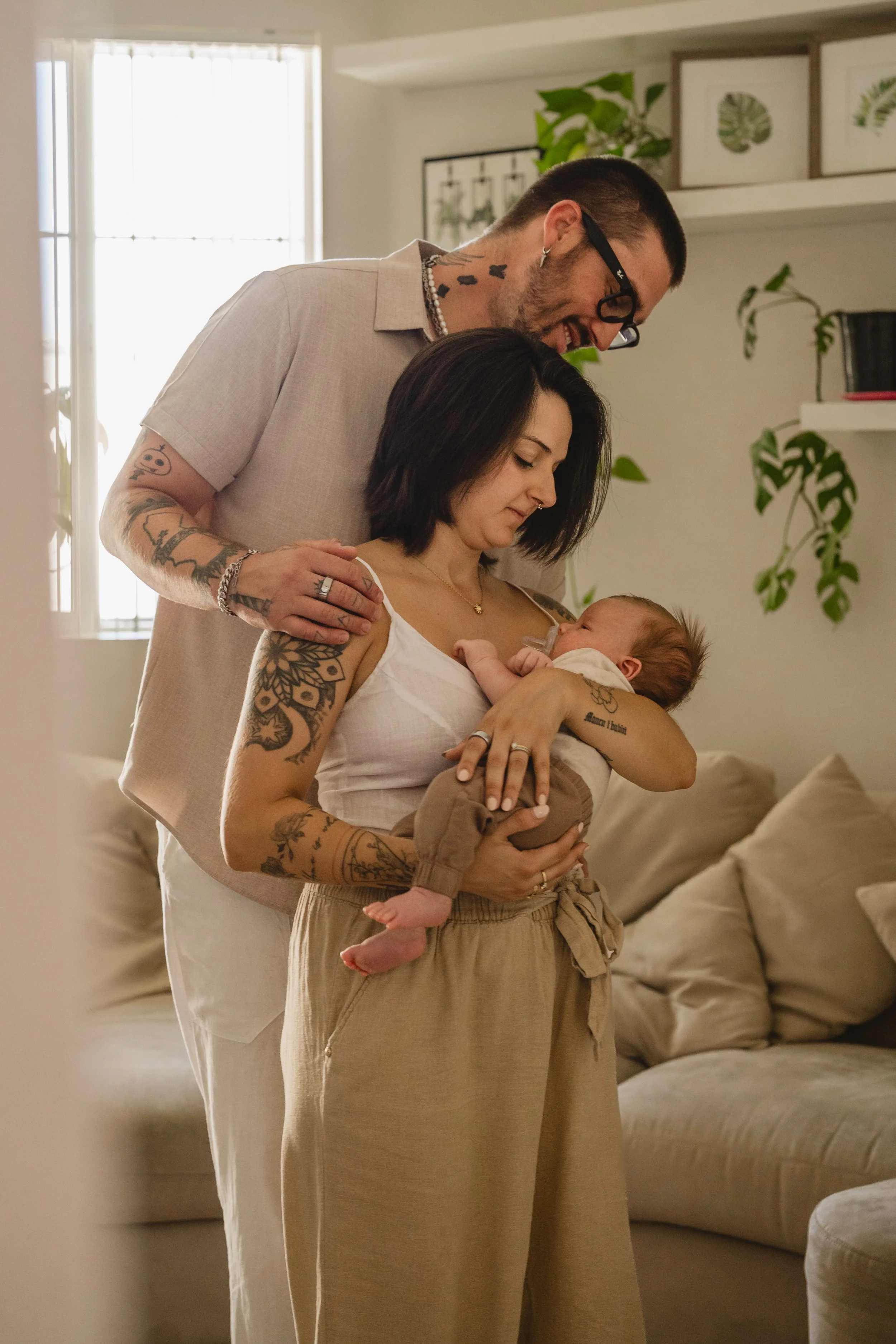 Standing in their living room, a father rests a hand on the mother's shoulder as both look down at the baby she holds, window light and houseplants filling the frame