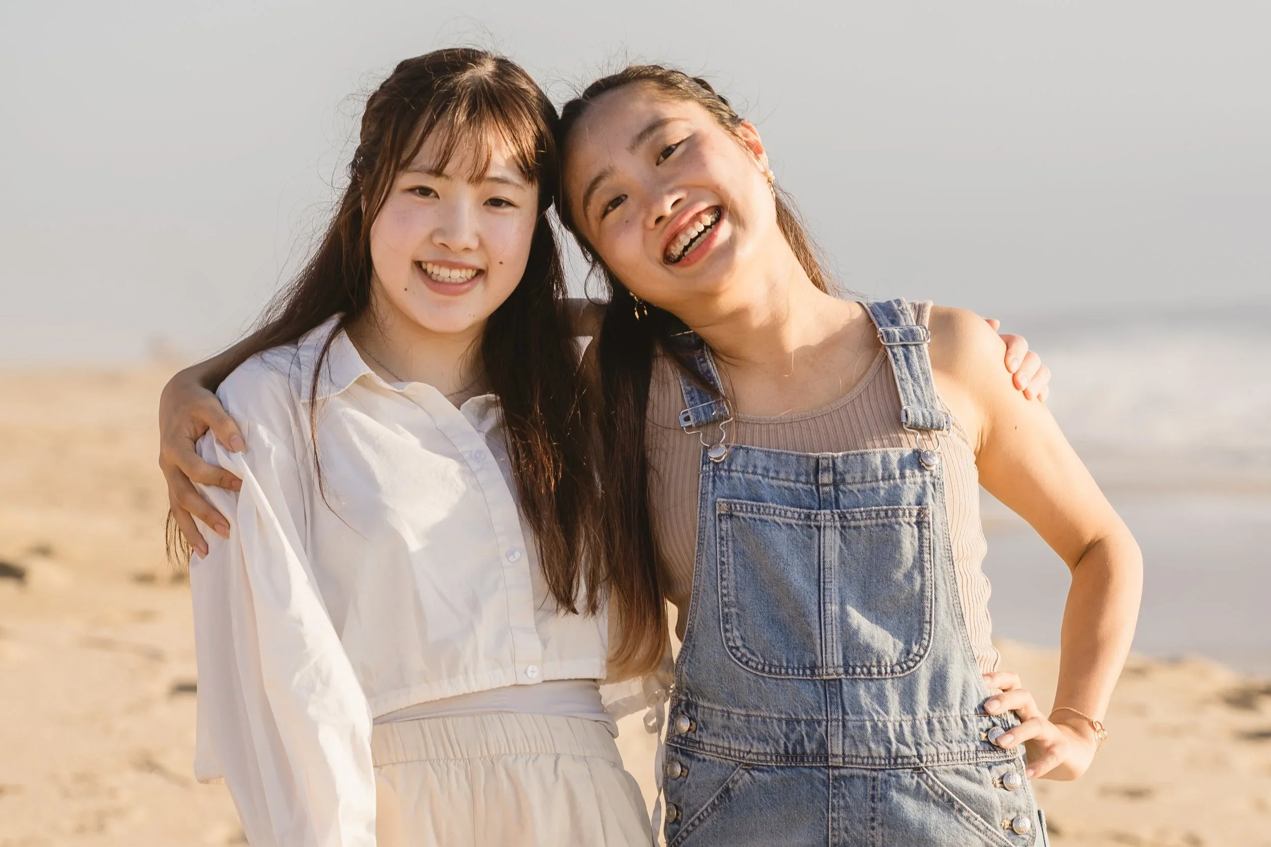 Two sisters stand with arms around each other's shoulders and heads tipped together, one in a white shirt dress and one in denim overalls