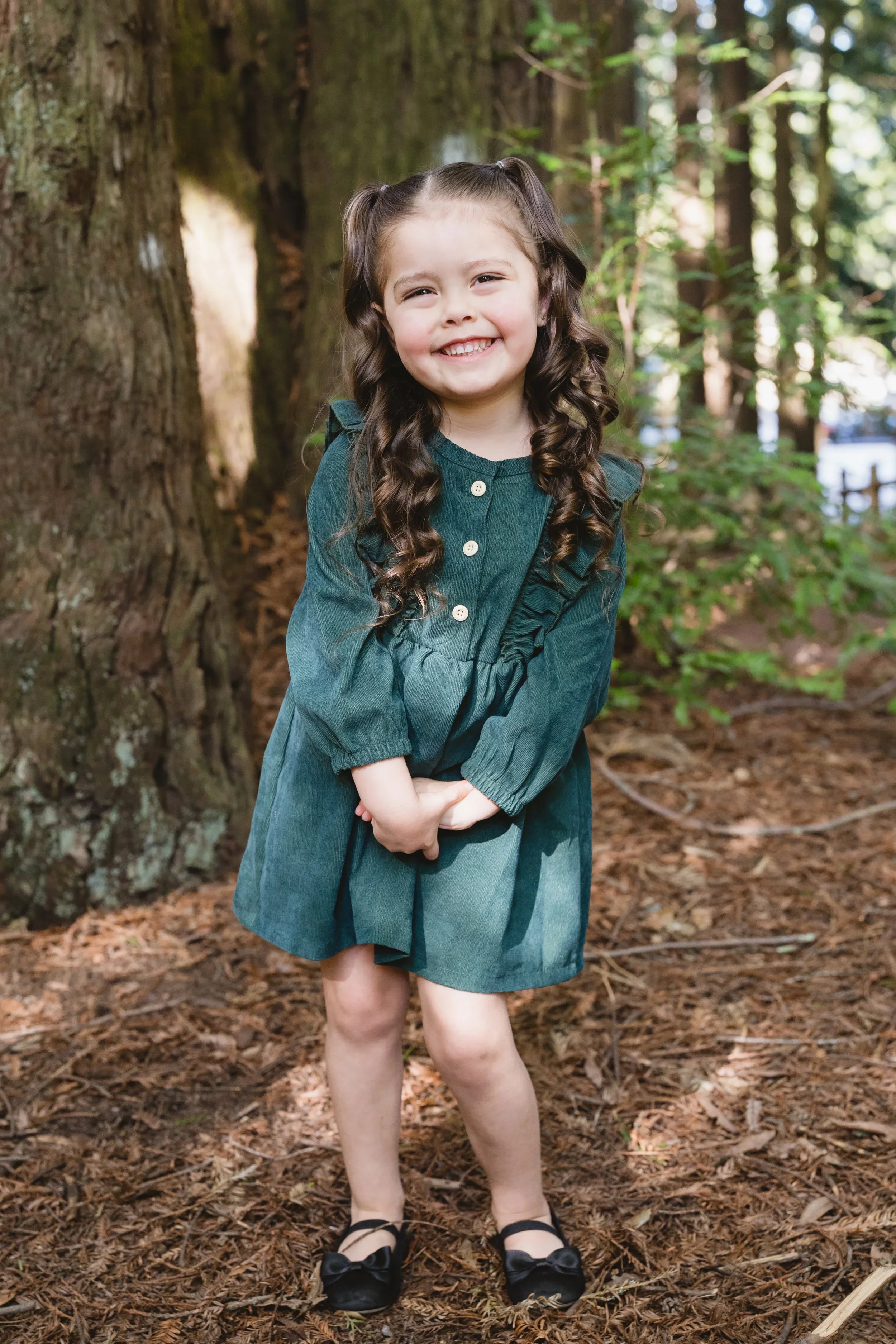 Young girl standing with her hands clasped in front of her, smiling toward the camera beside a redwood trunk on a pine-needle covered path