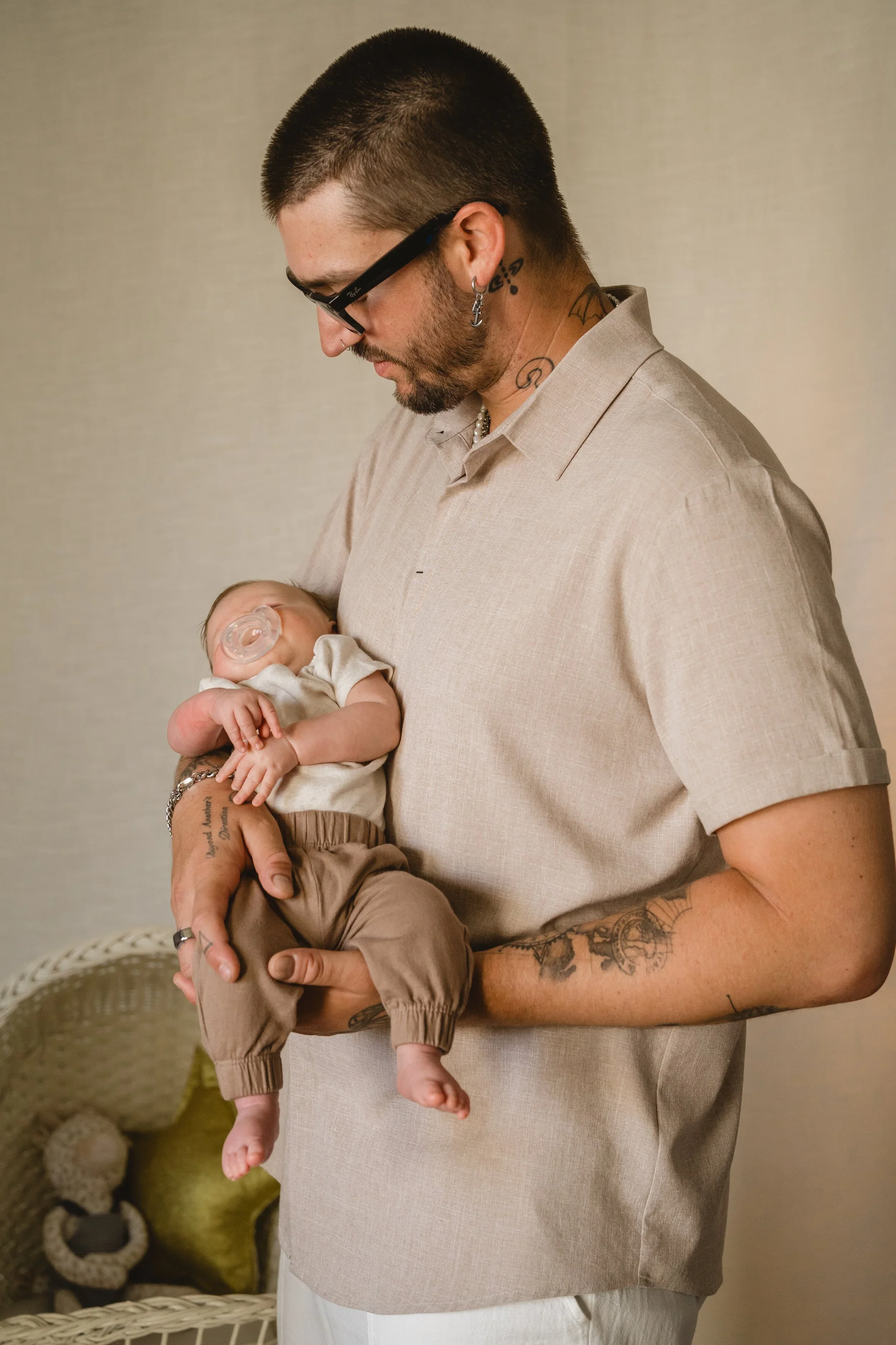 A father in a linen shirt looks down at the baby resting in the crook of his arm with a pacifier, a wicker bassinet just visible behind him