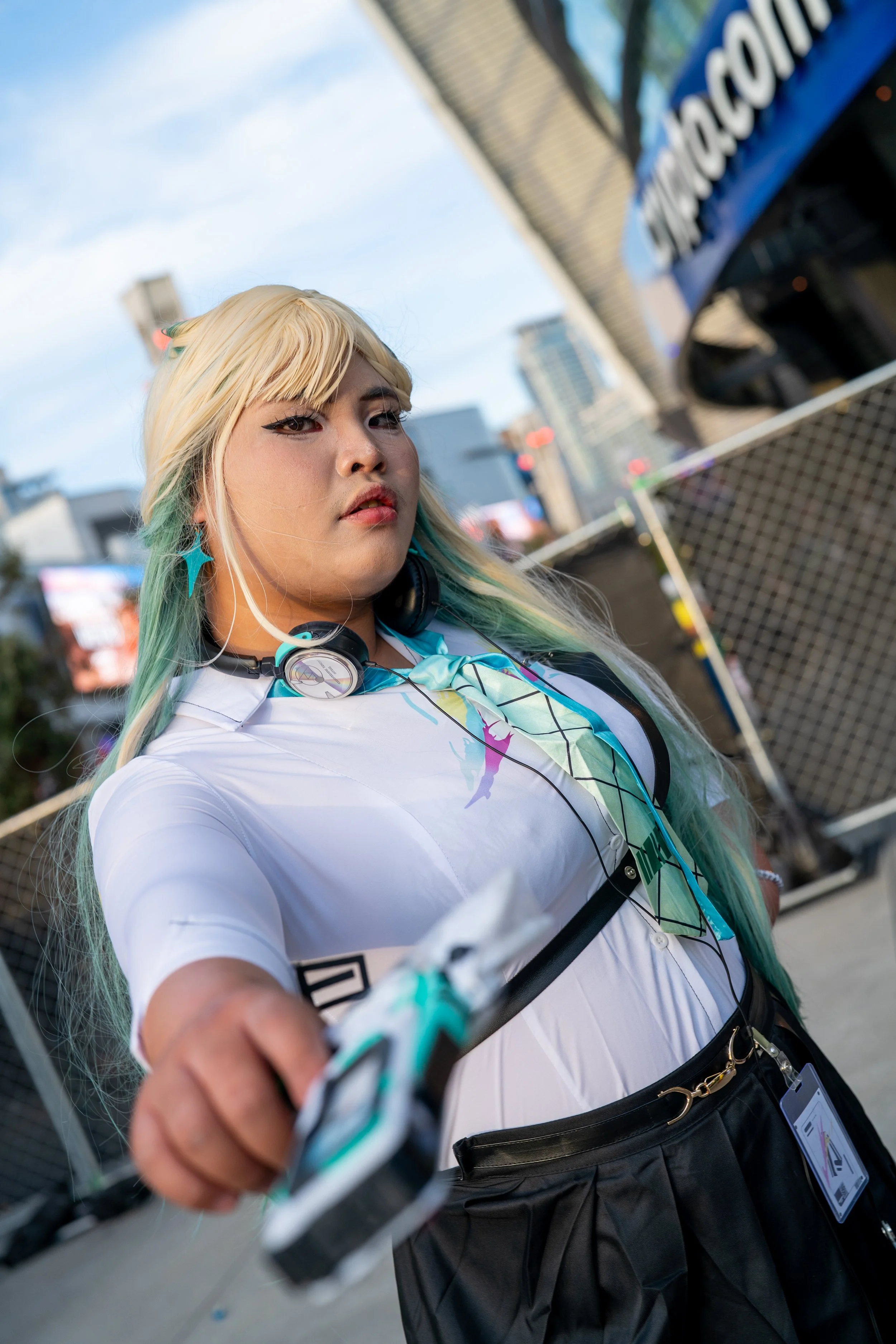 A person with long blonde hair and green tips wearing a white shirt, black skirt, and headphones around their neck. They are reaching out towards the camera with a cityscape background.