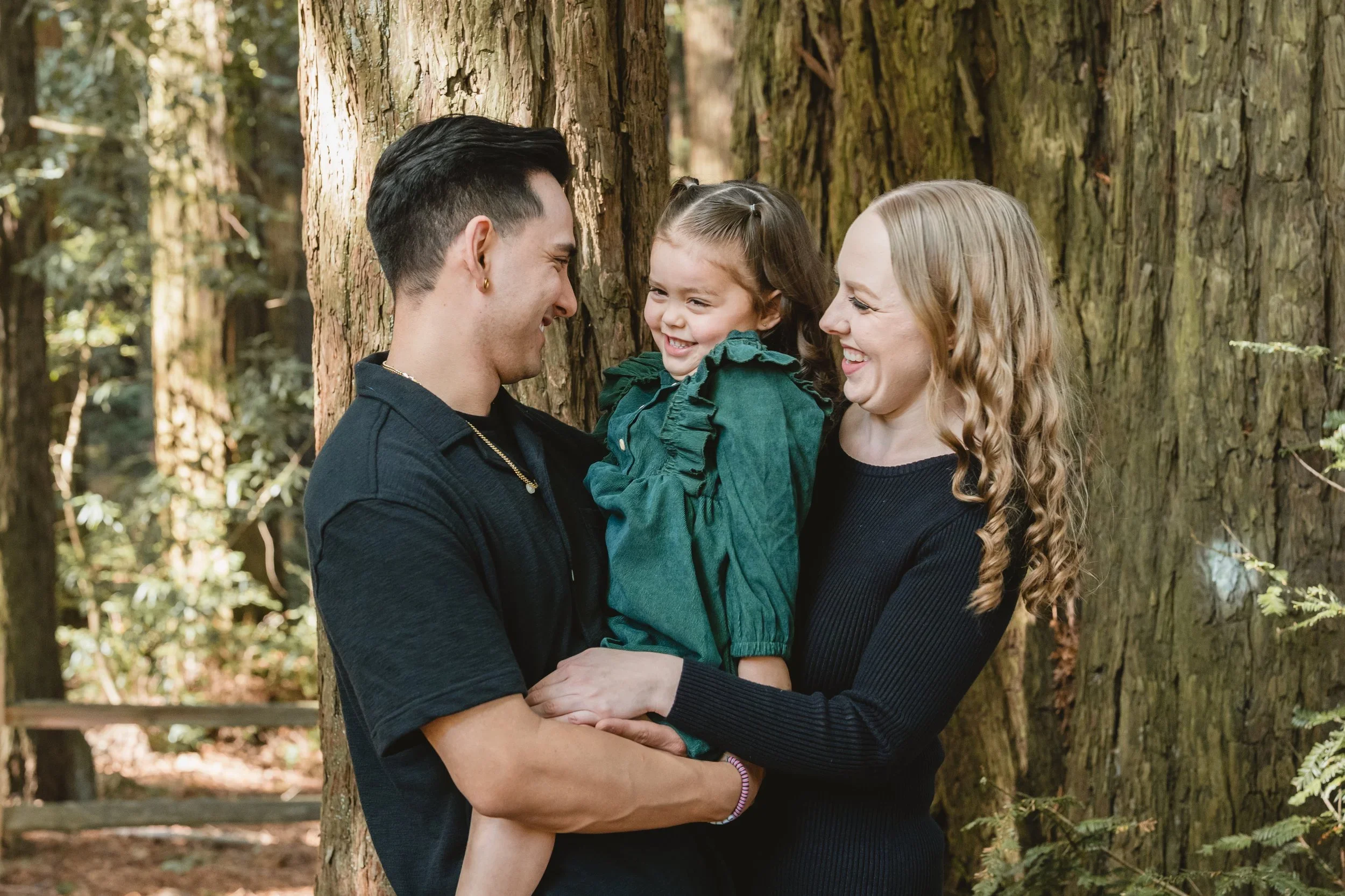 Parents hold their daughter between them at chest height, all three laughing as they look at each other in a grove of tall trees