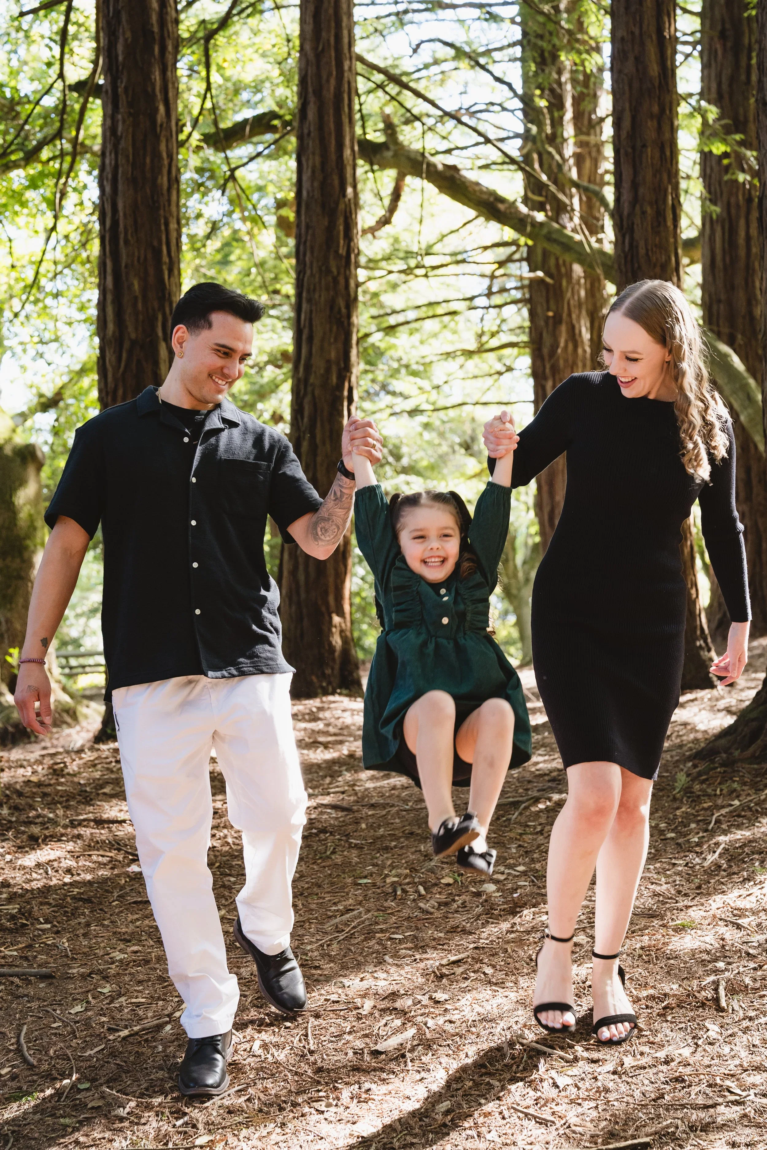 Mom and dad walk a shaded Oakland redwood trail holding their daughter's hands as she swings between them with her feet off the ground