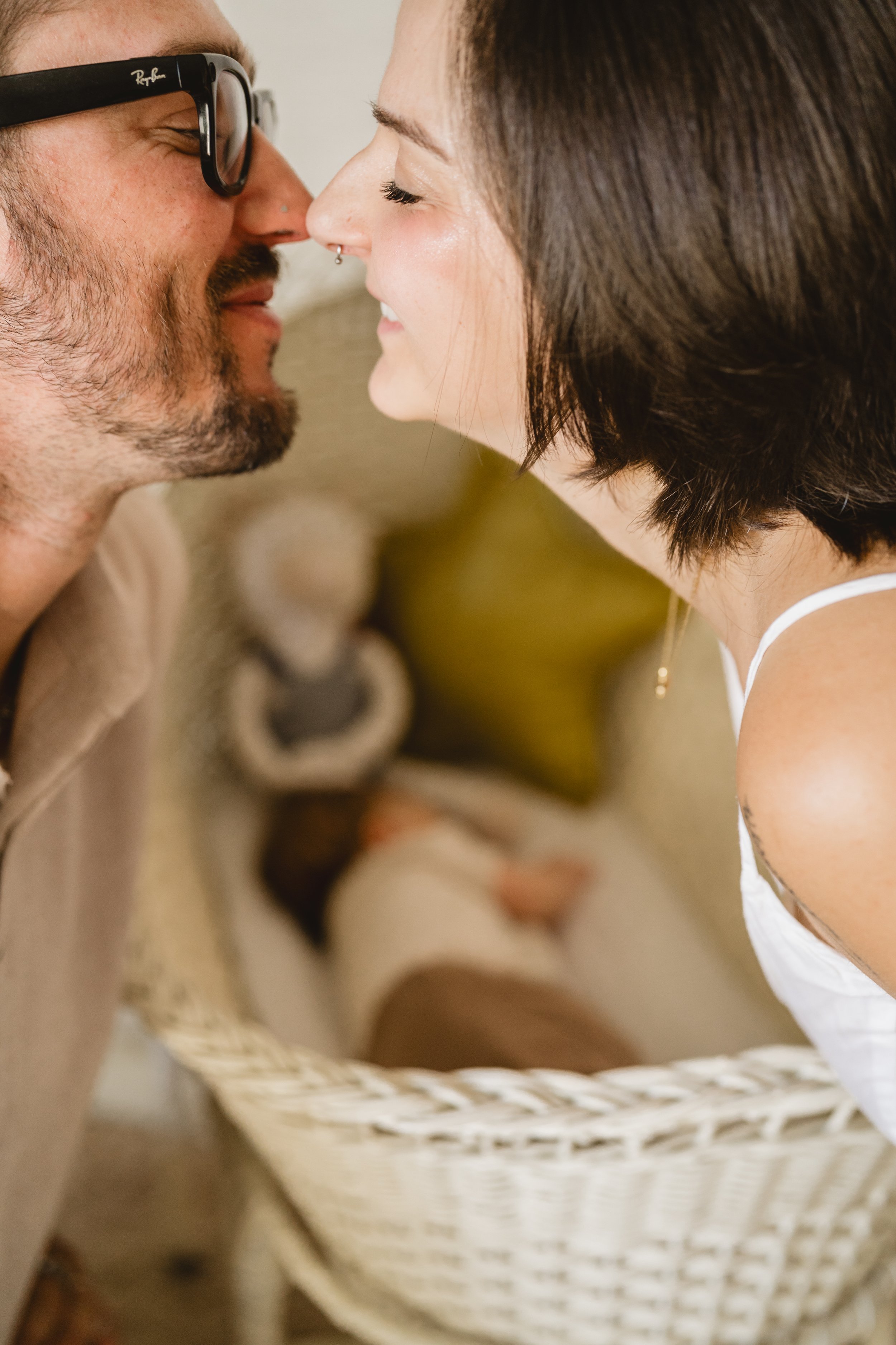 Close crop of two parents pressing their noses together and smiling, framed above the wicker bassinet where their baby rests out of focus below