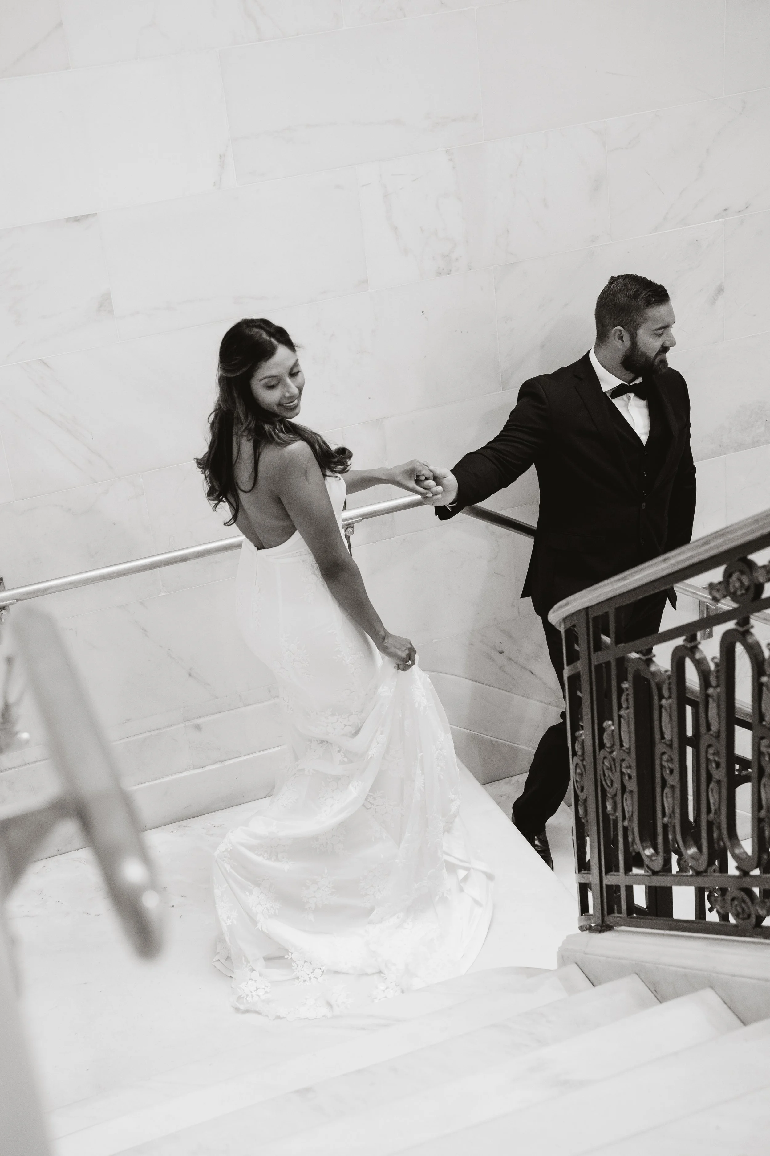 Black-and-white image of a bride glancing back over her shoulder while holding her train, walking down a staircase with her groom.