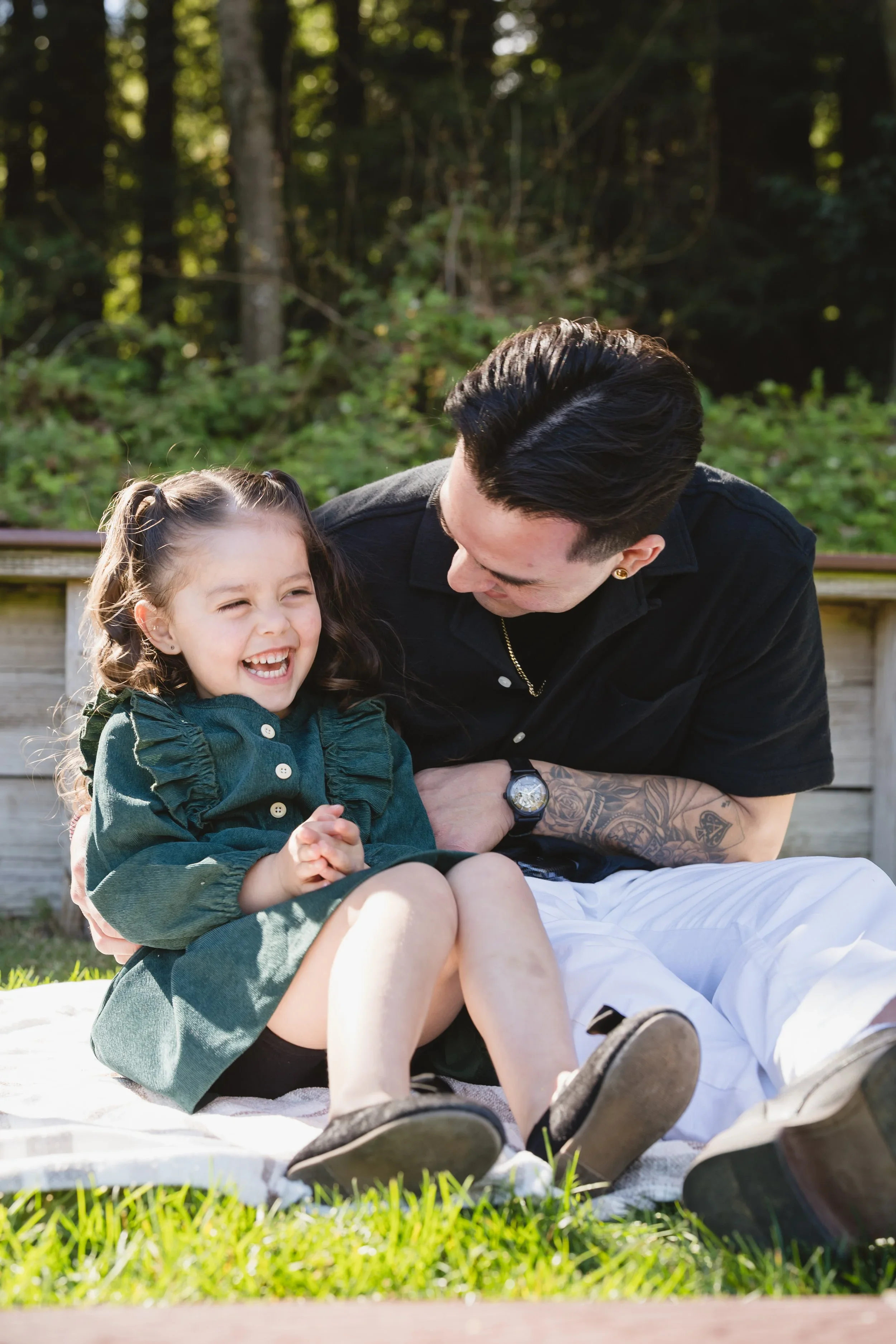 Girl in a green dress leans back into her dad's lap on a picnic blanket, laughing with her eyes squeezed shut while he looks down at her