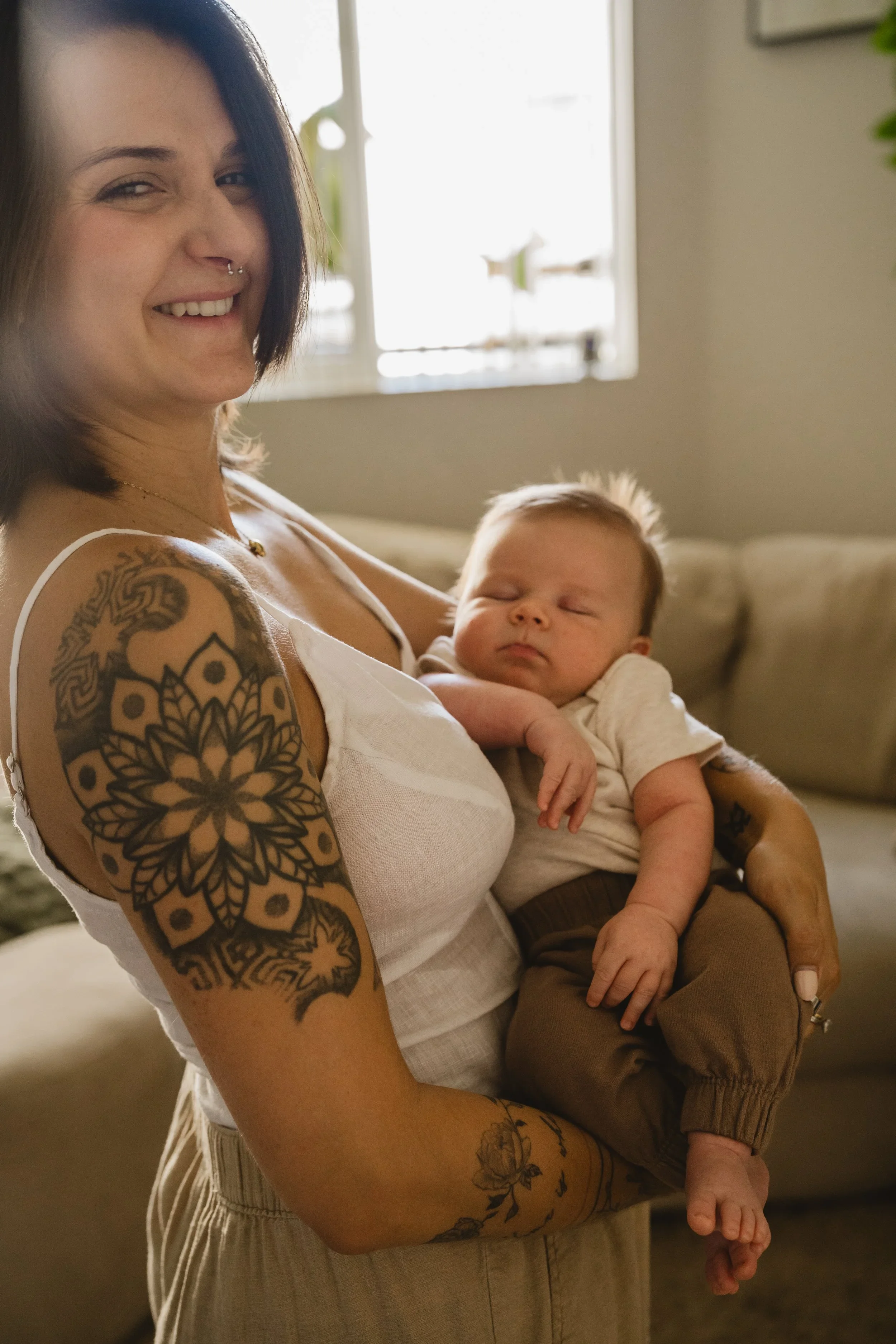 A mother turns toward the camera and smiles while holding her sleeping baby against her shoulder, backlit by a living room window
