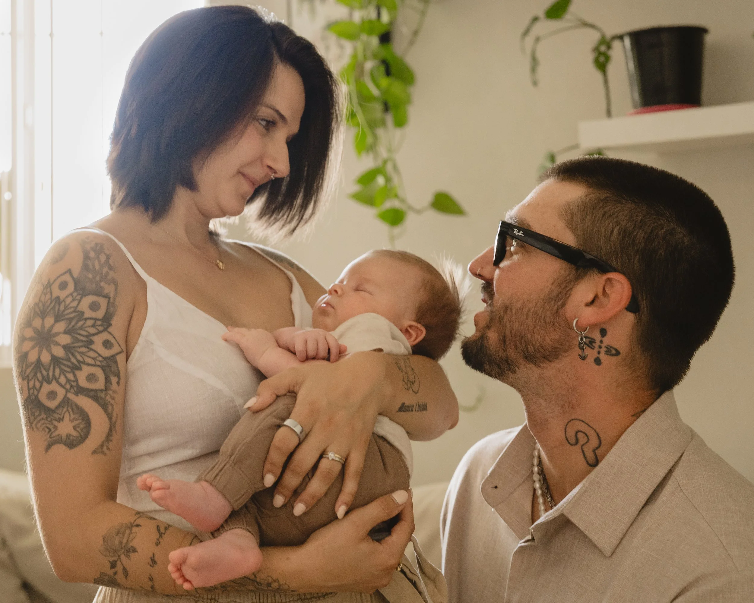 A mother holds her sleeping newborn while the father leans in from the right, the two parents facing each other with the baby between them