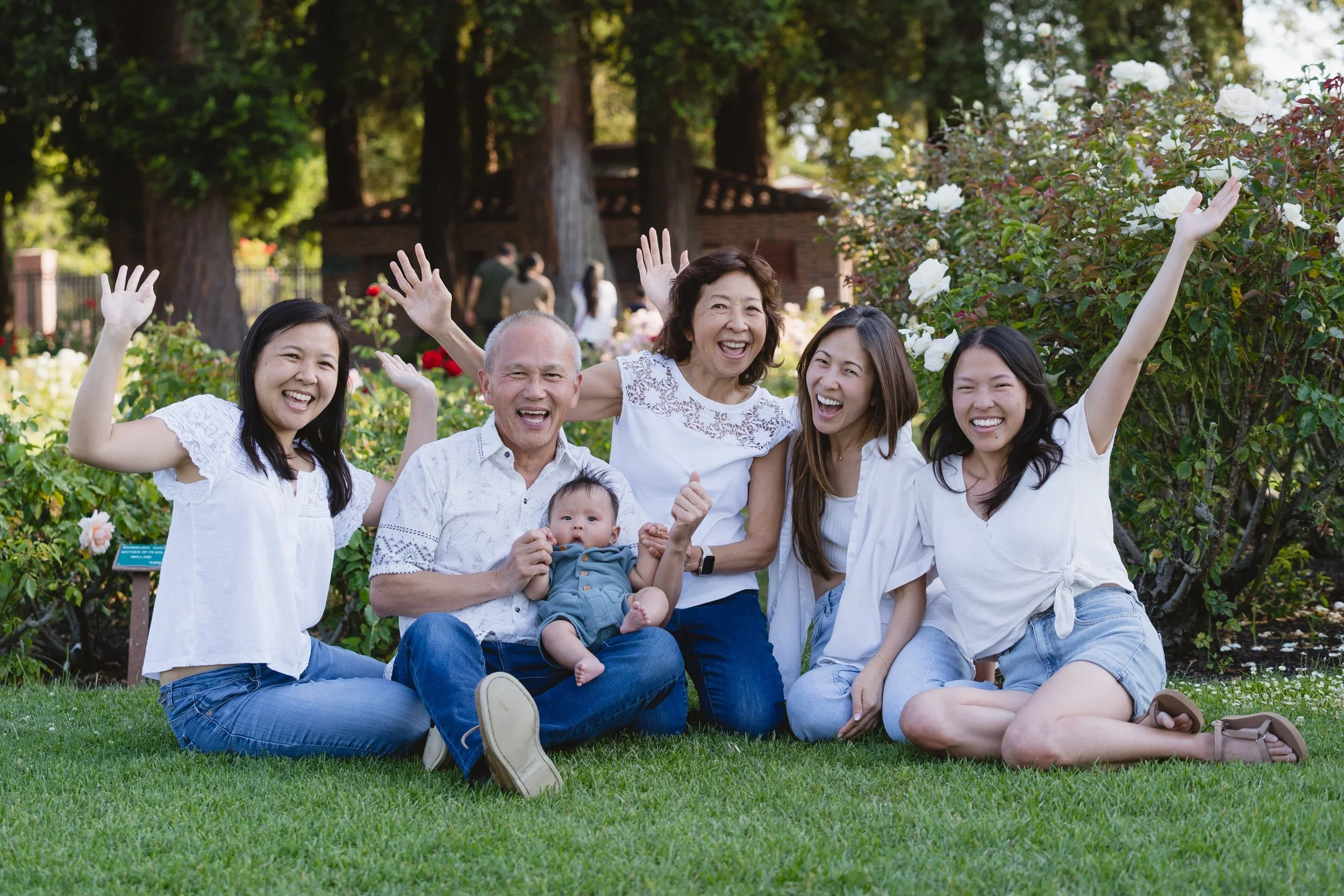 Six family members sit together on the lawn with their arms thrown up, grandfather holding the baby on his lap, beside a white rose bush at the San Jose Rose Garden.
