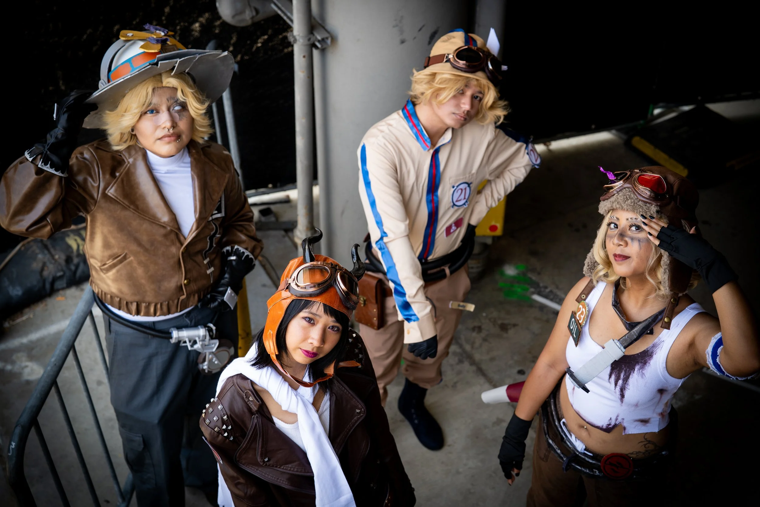 Group of five young people dressed in steampunk costumes with goggles, leather and vintage clothing, standing and sitting in an industrial setting.