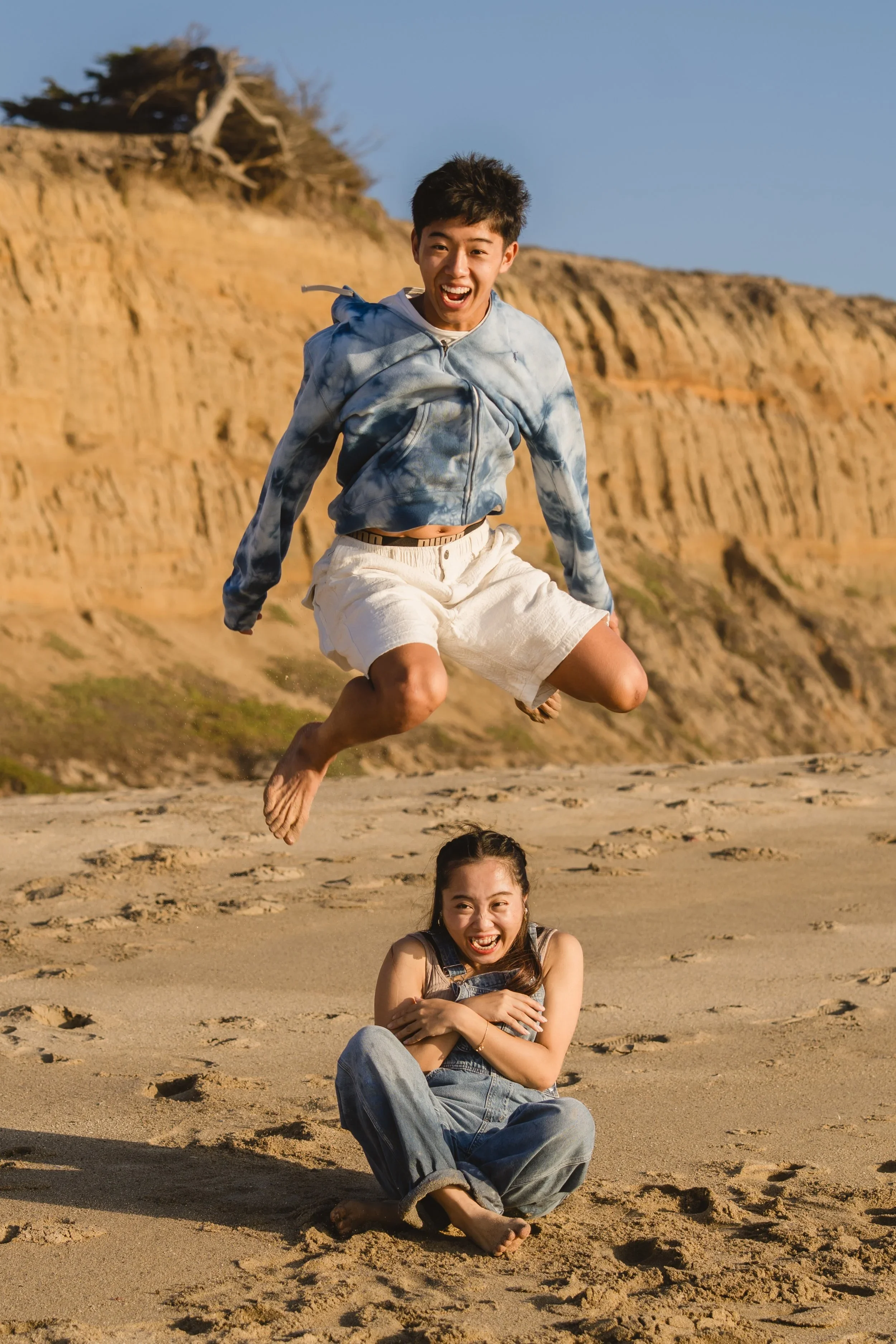 A young man leaps mid-air above his sister, who sits cross-legged on the sand laughing, with a cypress-topped bluff behind them
