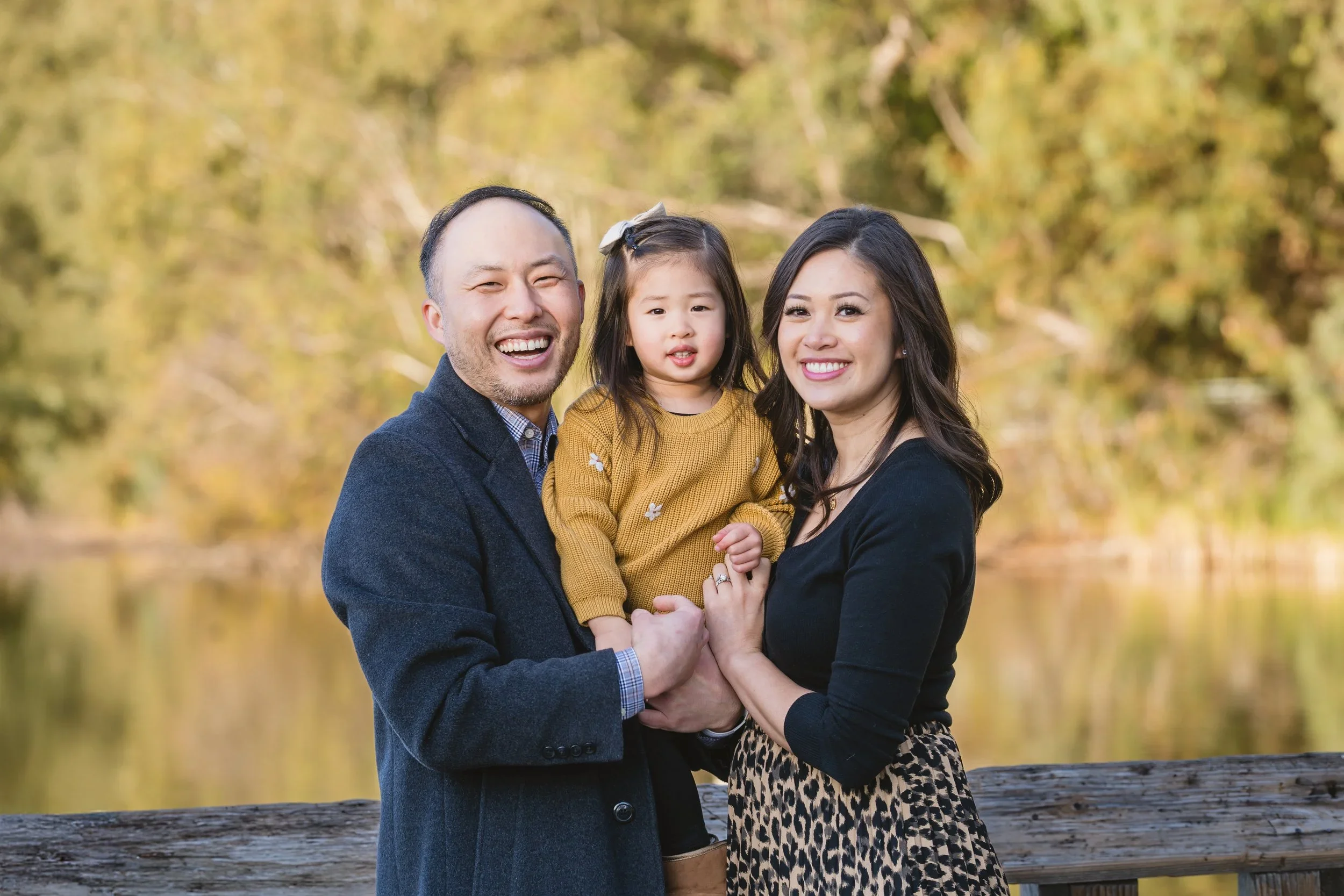 Mom, dad, and their daughter stand at a wooden railing at Vasona Lake, smiling directly at the camera with golden autumn trees softly blurred behind them.