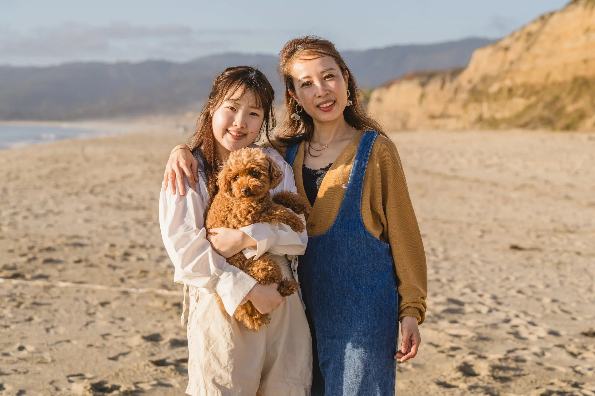 A daughter cradles the family's curly poodle mix while her mom keeps an arm around her, sandstone bluffs and open beach stretching behind