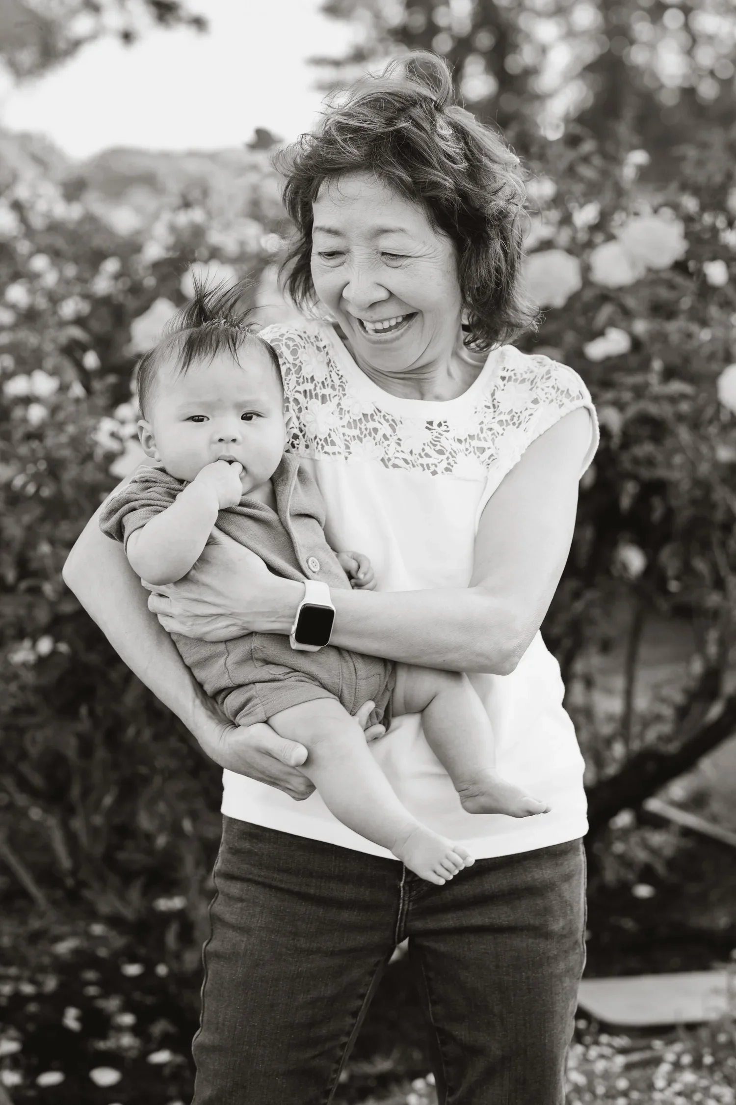 Grandmother in a white lace top carries her infant grandson on her hip and laughs down at him, photographed in black and white among the rose beds.