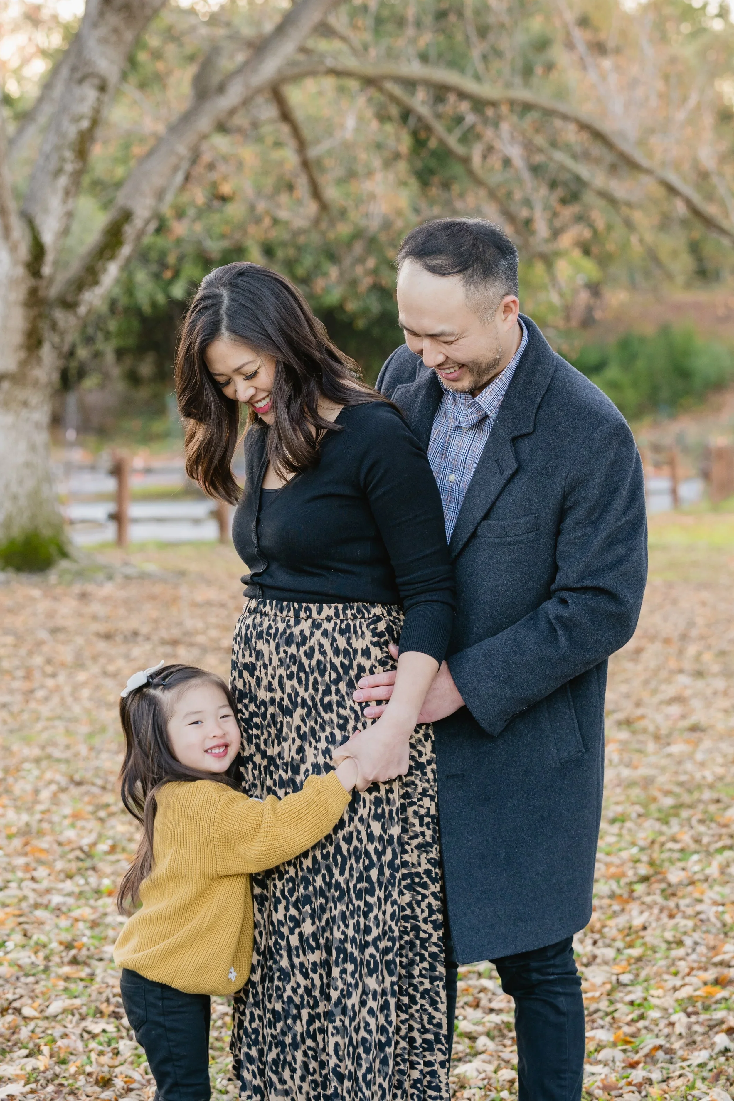 Dad cradles mom's pregnant belly with both hands while their daughter reaches in to join, all three smiling down under bare autumn branches.