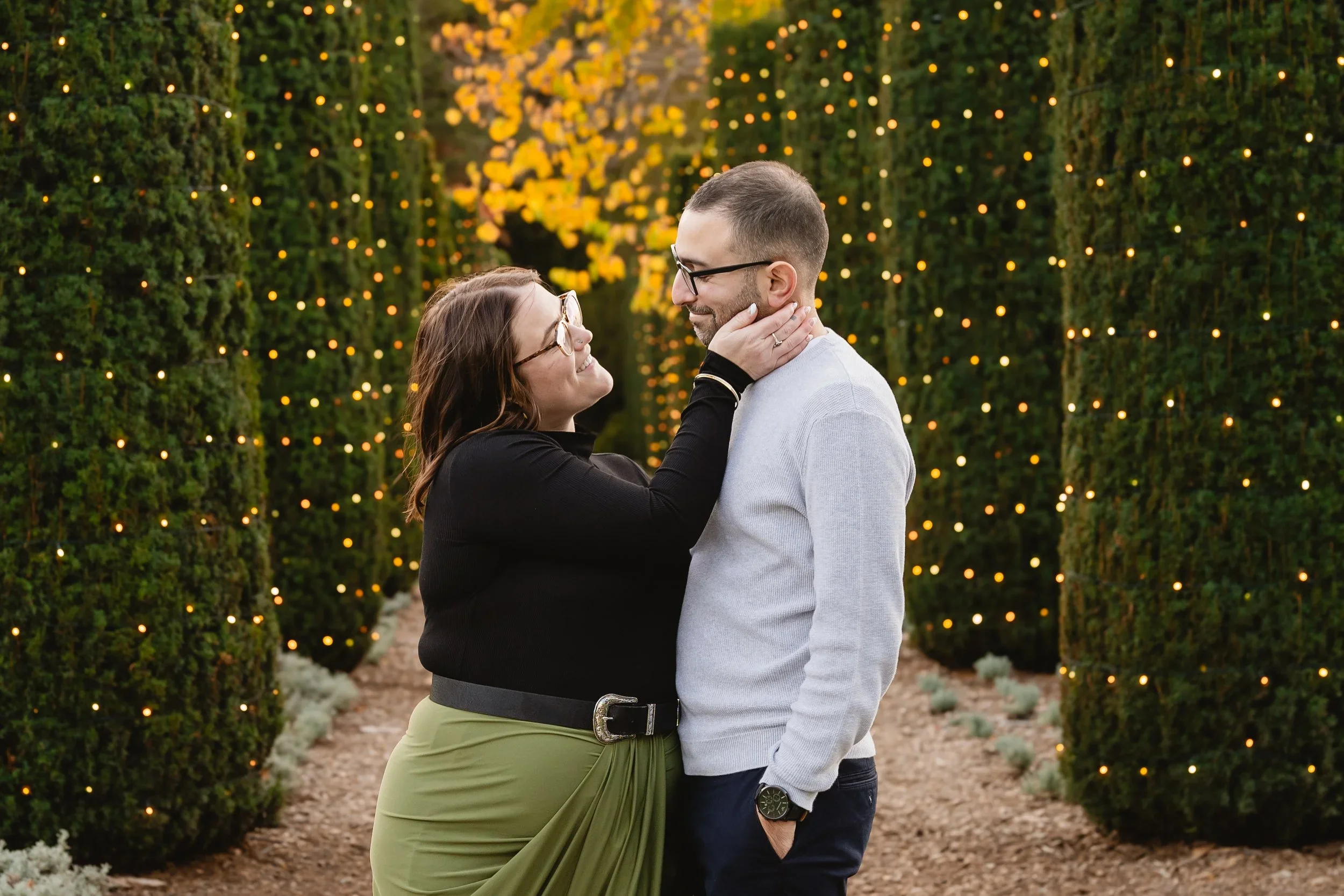 A woman reaches up to touch a man's face on a hedge-lined path strung with warm string lights during a winter engagement session at Filoli.