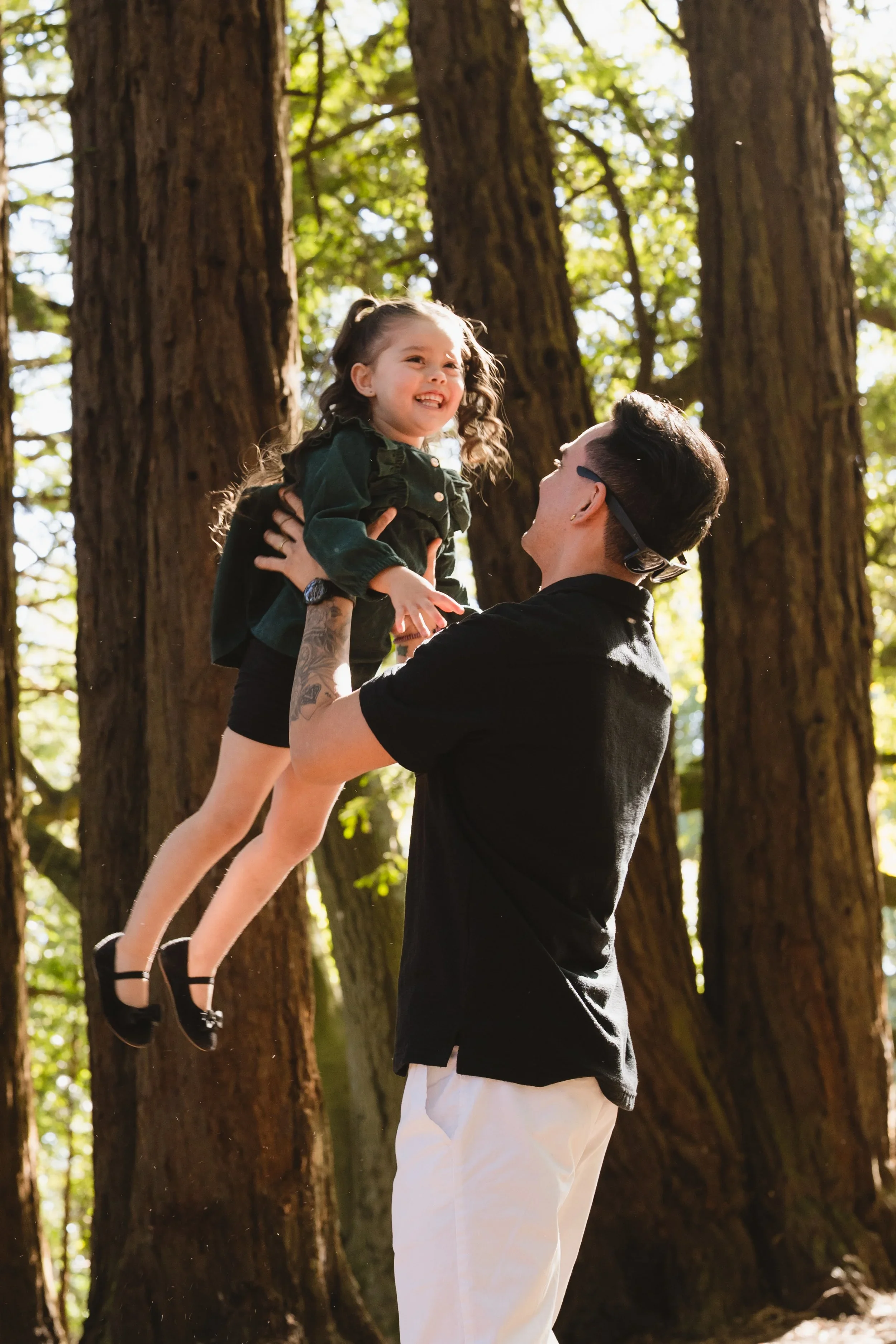 A father holds his daughter high in the air between towering trunks, both grinning at each other with sunlight coming through the canopy behind them