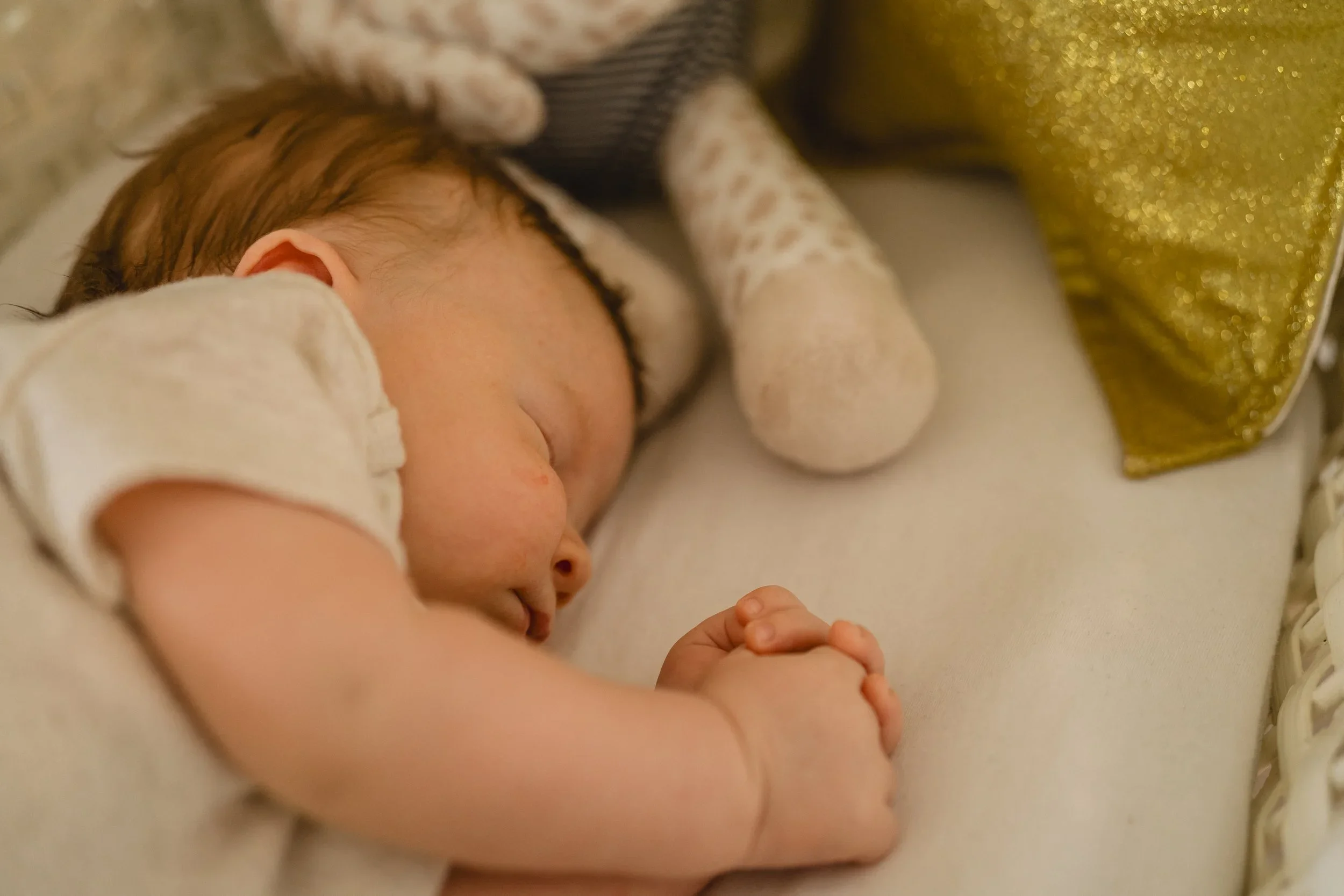 Overhead close-up of a newborn asleep on a cream bassinet sheet with hands curled together, a knit toy and gold sequin star pillow tucked alongside