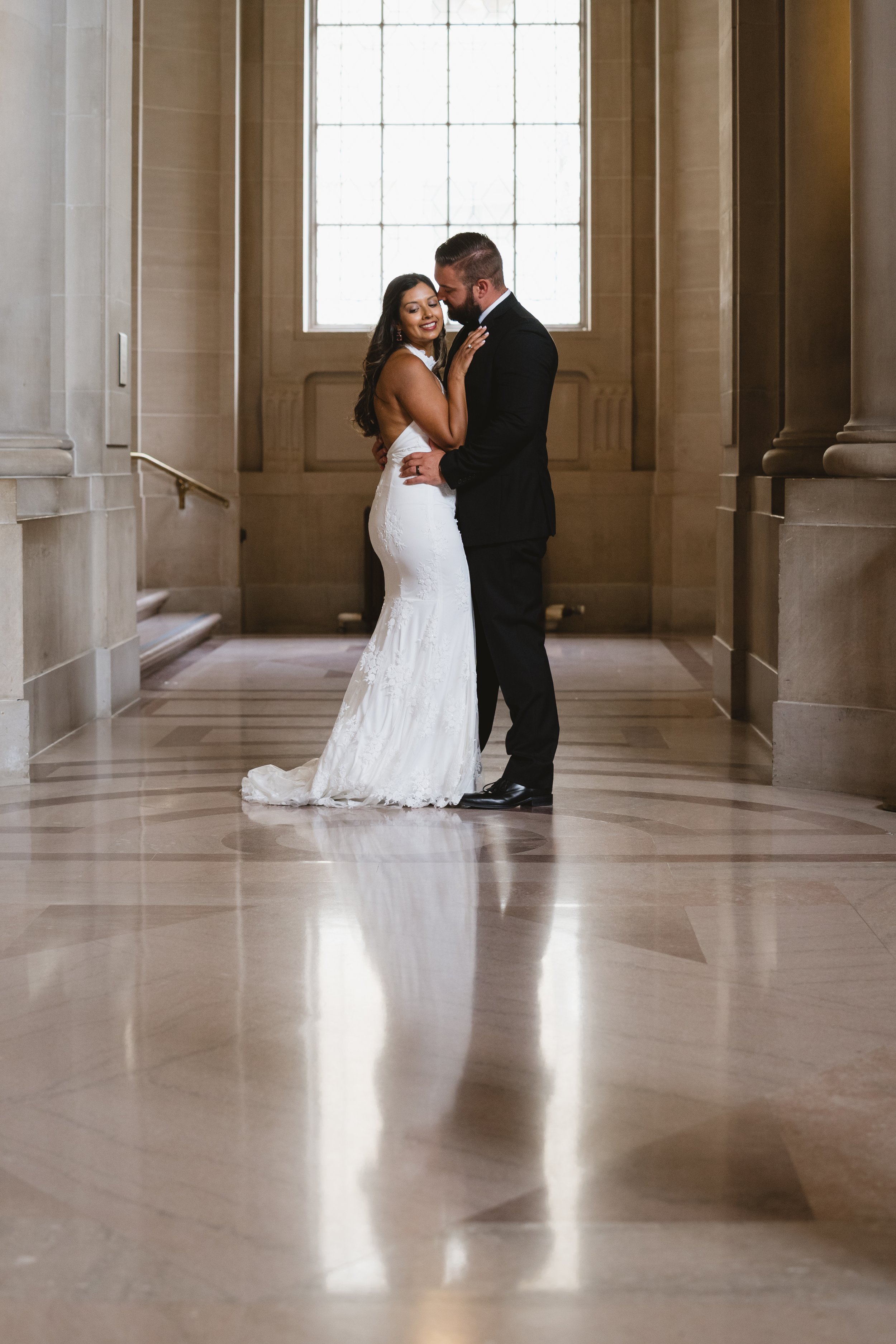 Couple embracing near a tall leaded window in a marble corridor, their reflection visible on the polished floor.