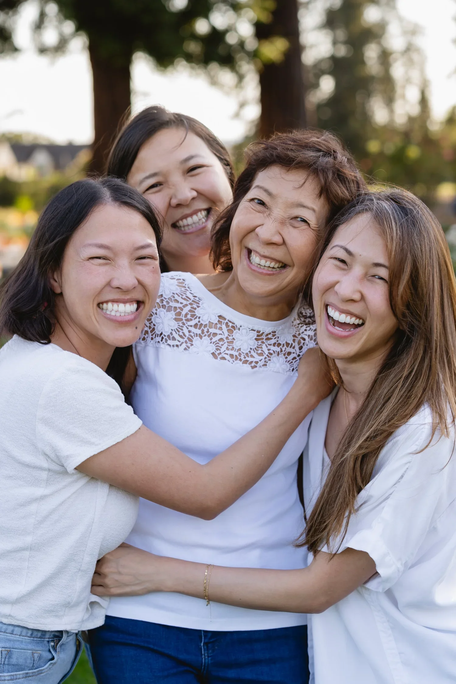 Close crop of three daughters squeezing in around their mom mid-laugh, all in white tops, with blurred trees and rose beds behind them.