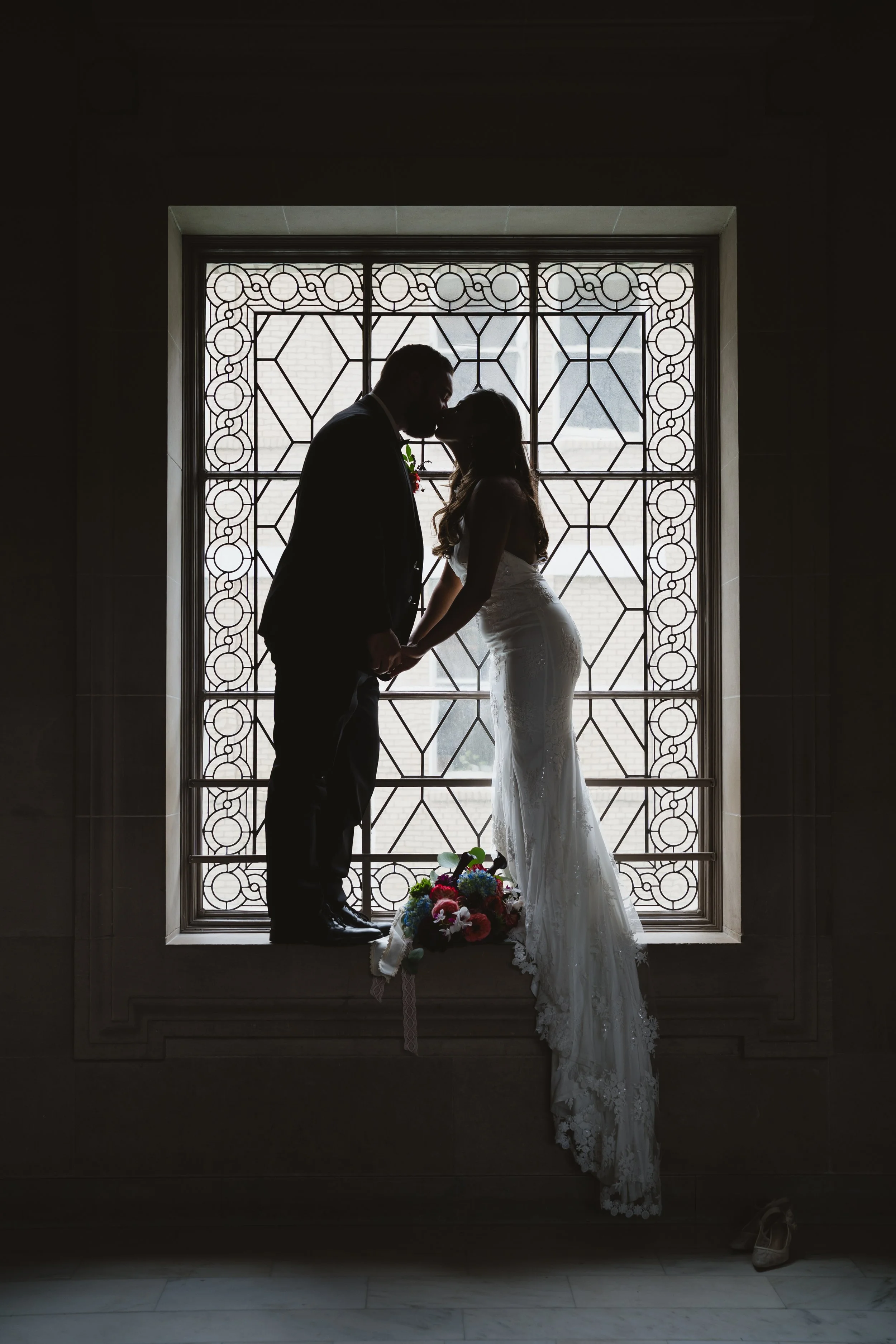 Silhouette of a bride and groom kissing in front of a decorative window, with a bouquet of flowers placed on the windowsill and one shoe on the floor.