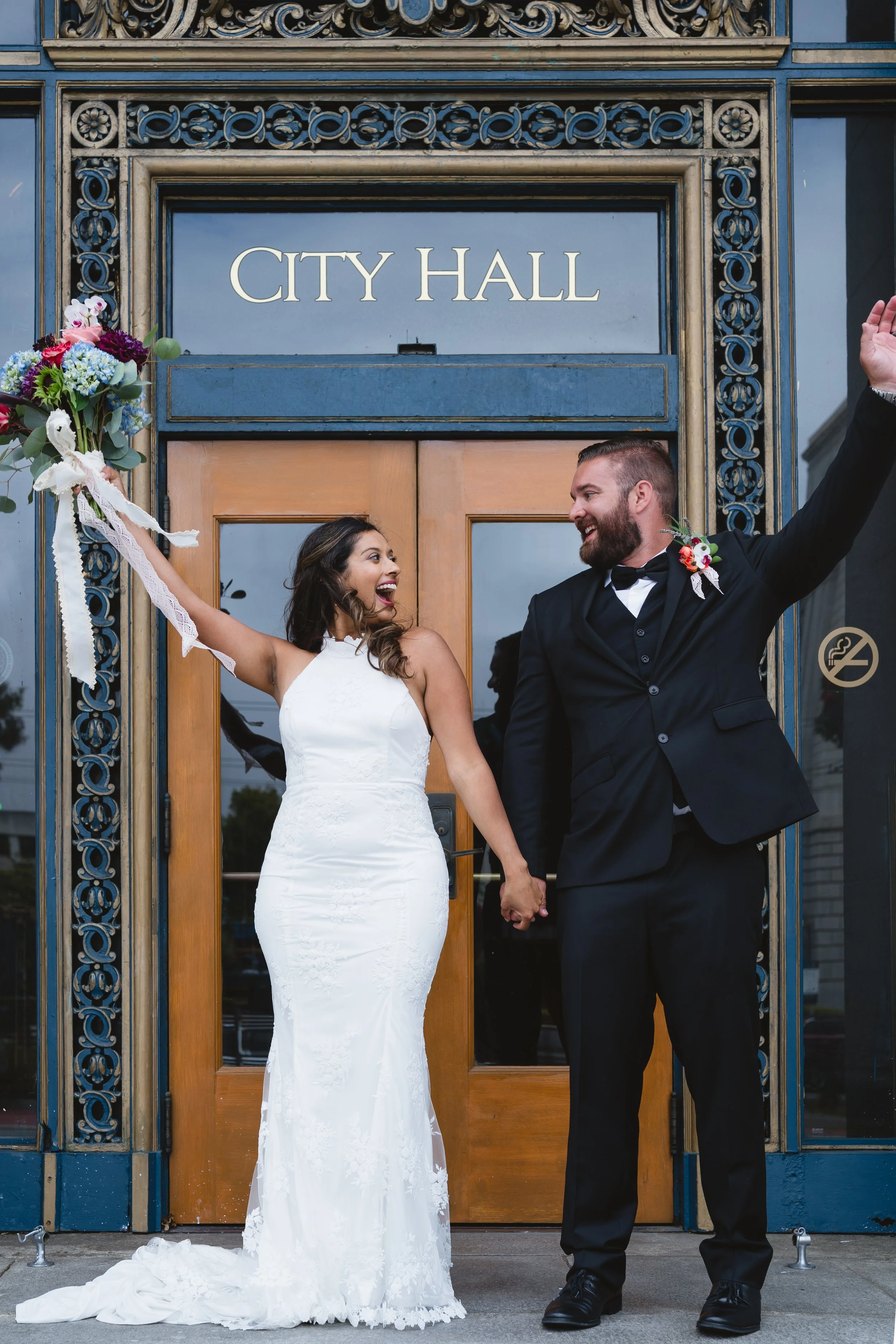Bride and groom cheering with arms raised and hands clasped outside the City Hall doors.