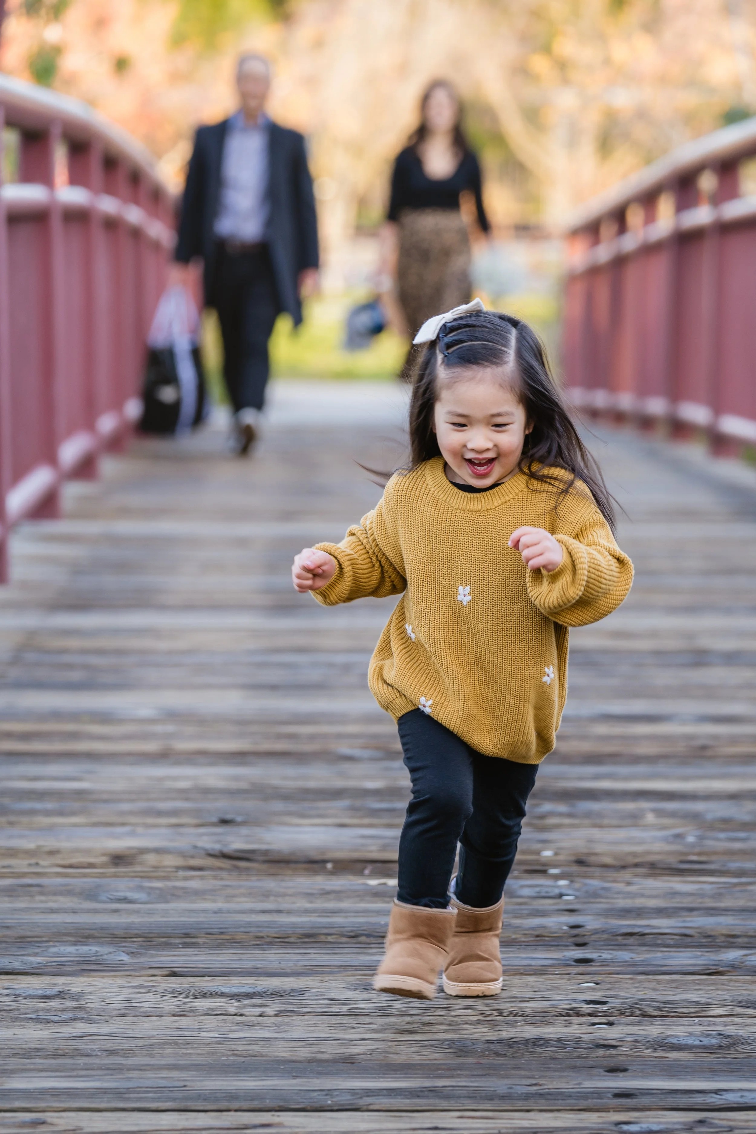 A young girl running and smiling on a wooden bridge during autumn, with blurred adults walking behind her in the background.