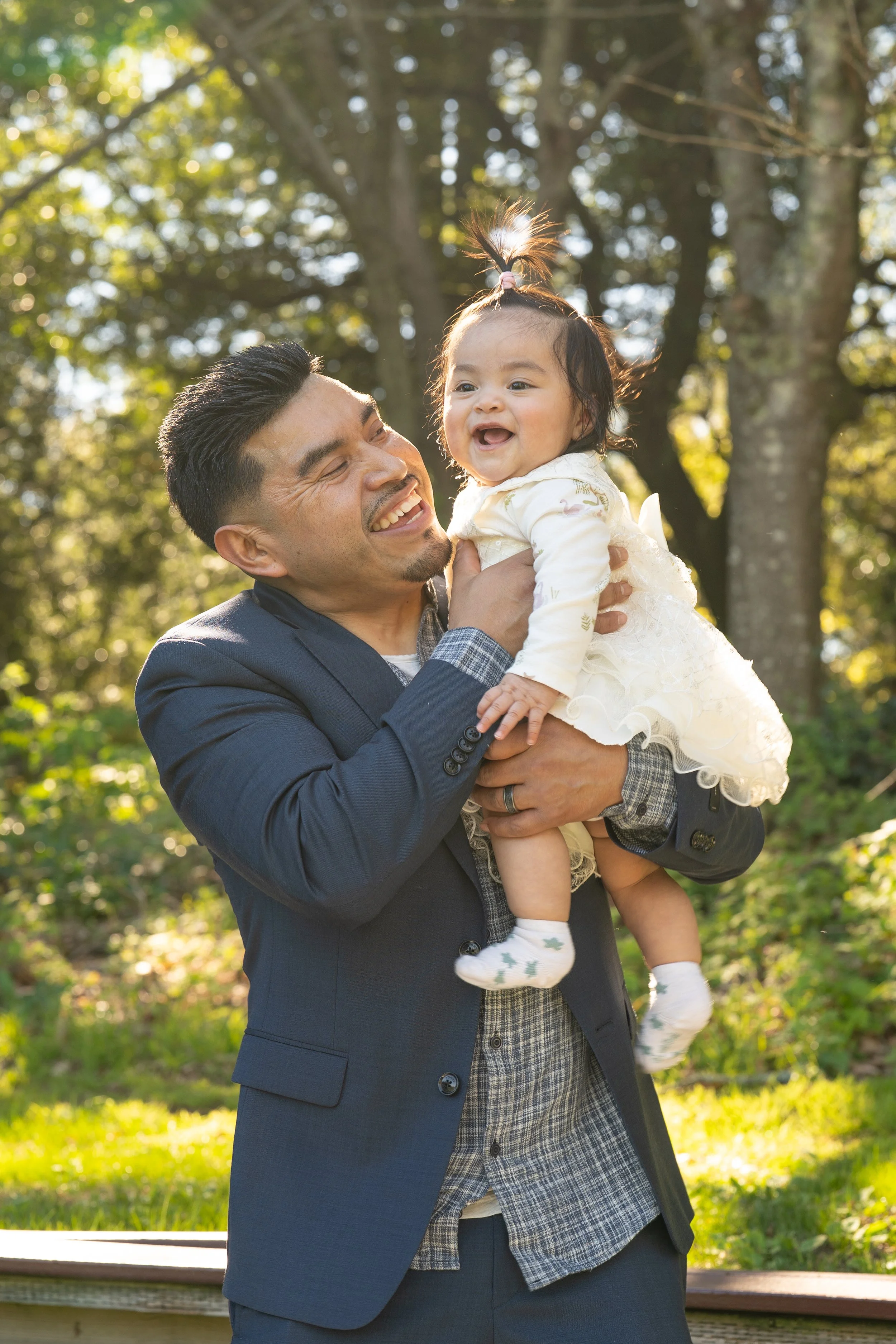 A man in a suit holding a smiling toddler girl outdoors in a park with trees and sunlight in the background.