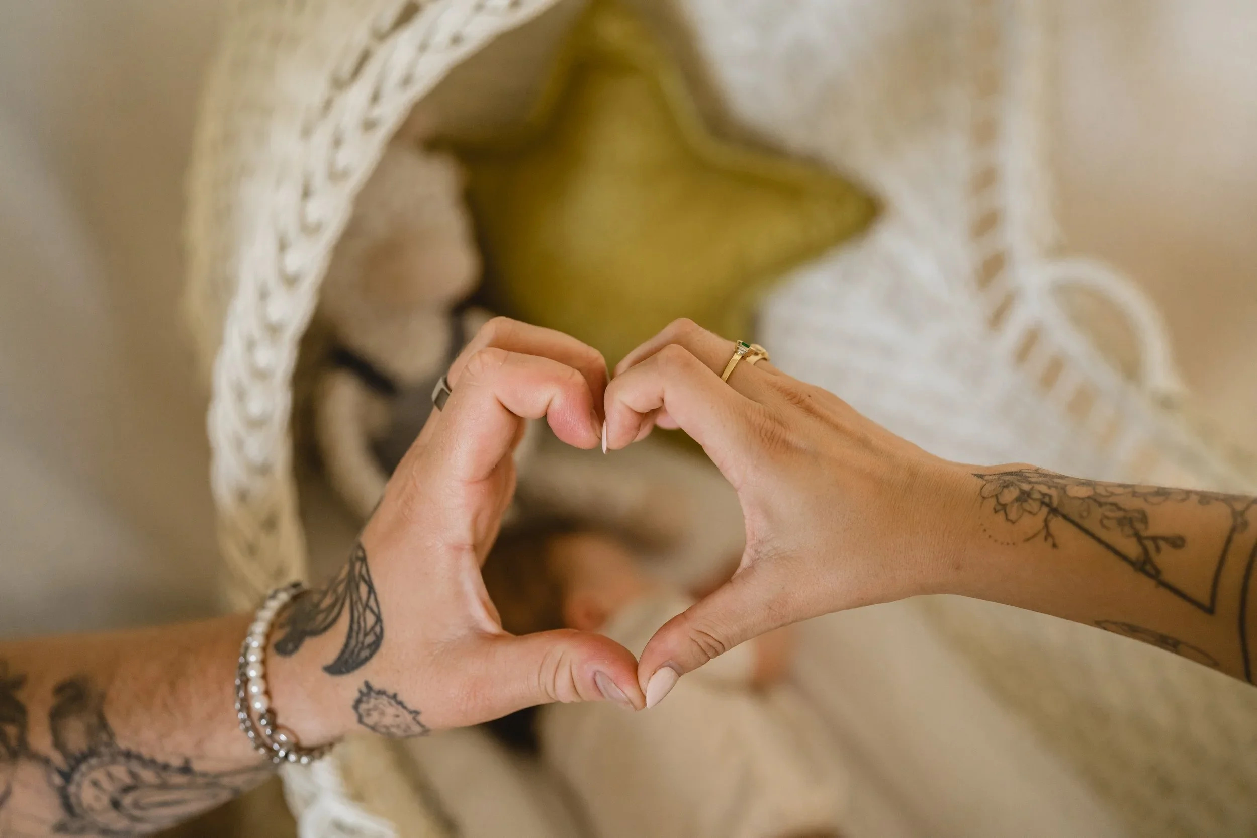 Two tattooed hands meet to form a heart shape above a wicker bassinet, the sleeping baby and a gold star pillow soft in the background