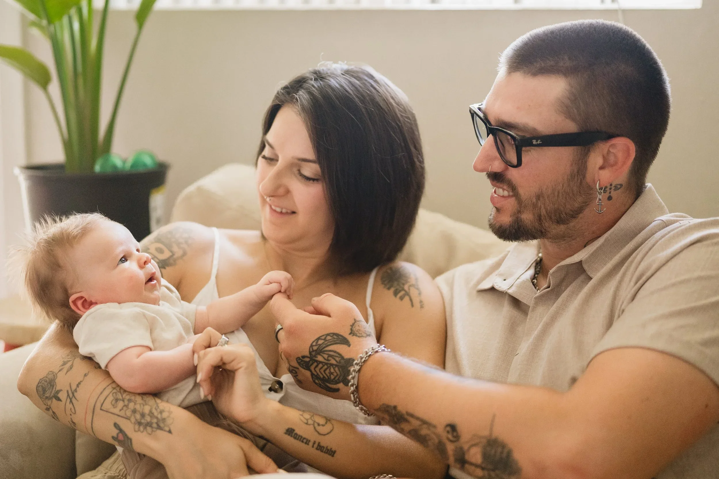 Both parents lean in and smile as their wide-awake baby lies in mom's arms and grips her finger, seated together on a sofa beside a houseplant