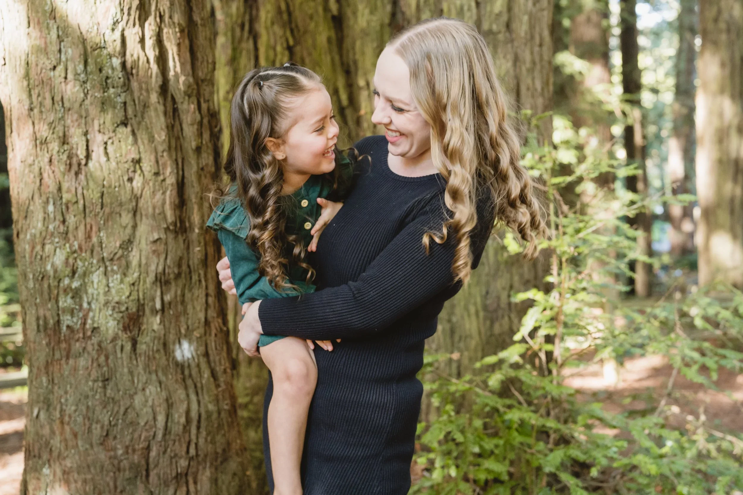 A mom carries her daughter on her hip beside a wide redwood trunk, the two turned toward each other and smiling in dappled afternoon light