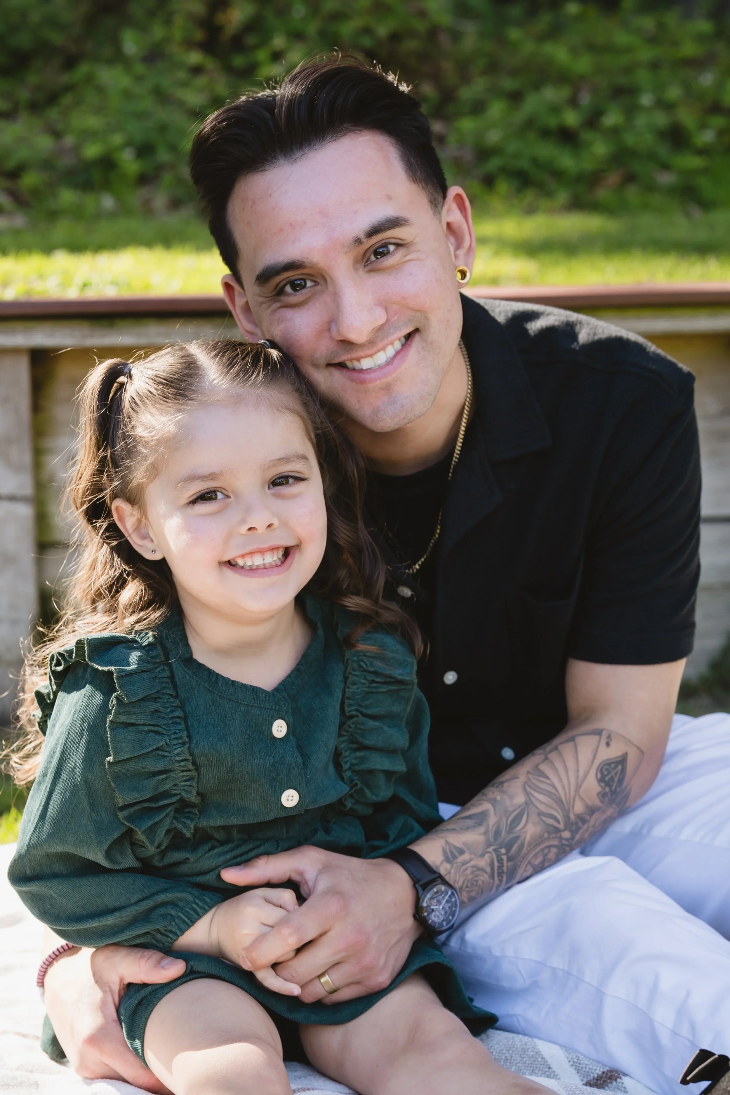 Dad sits with his arms around his daughter on a blanket, his head close to hers, the two of them smiling directly at the camera in afternoon sun