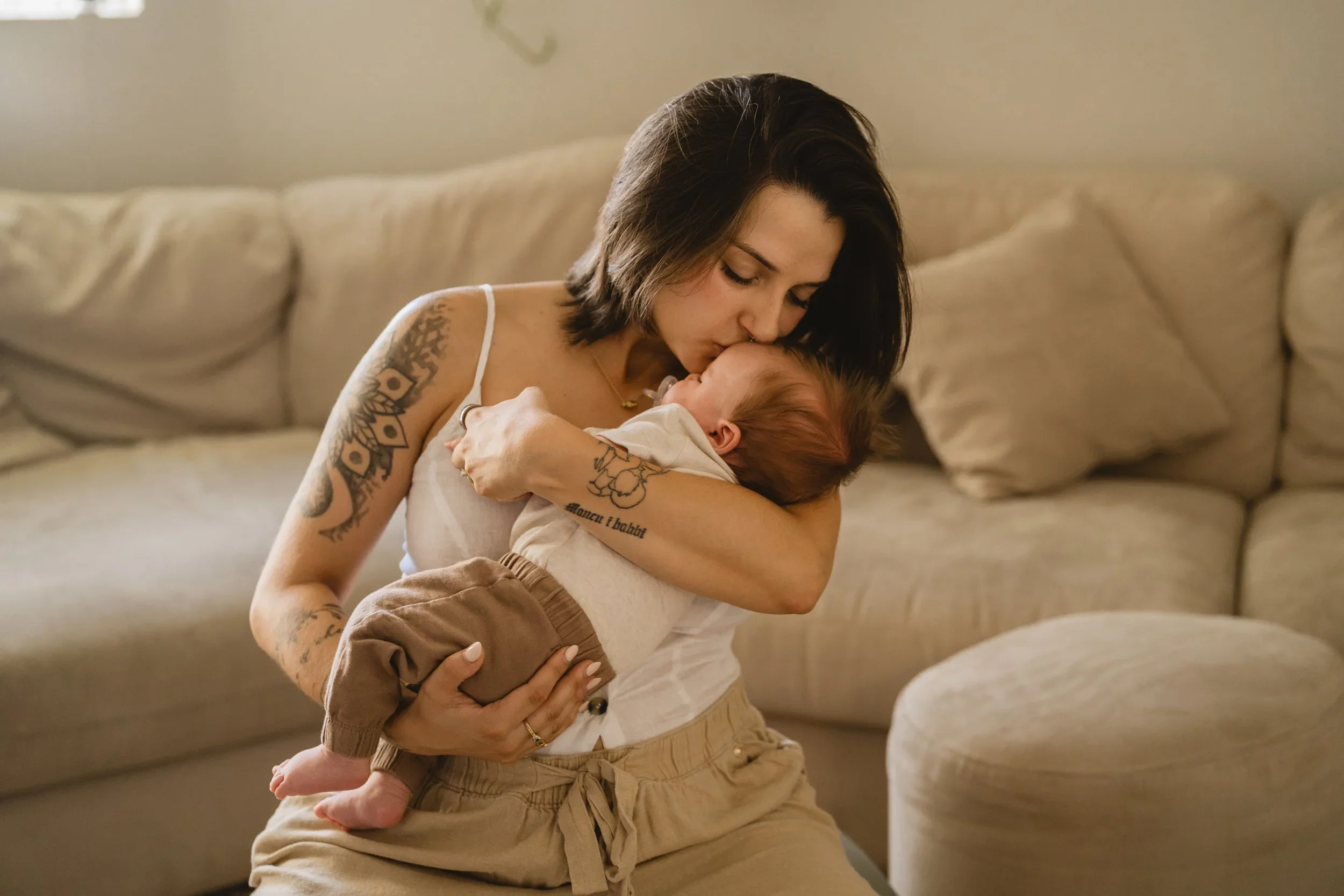 Seated on a linen sofa, a mother with tattooed arms kisses the top of her newborn's head while holding the baby upright against her chest