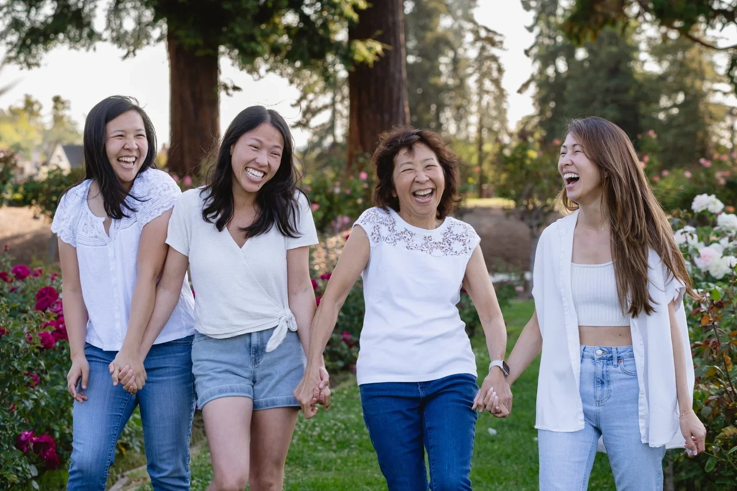 A mom and her three grown daughters walk toward the camera hand in hand, all laughing, past blooming rose bushes and tall redwoods in San Jose.