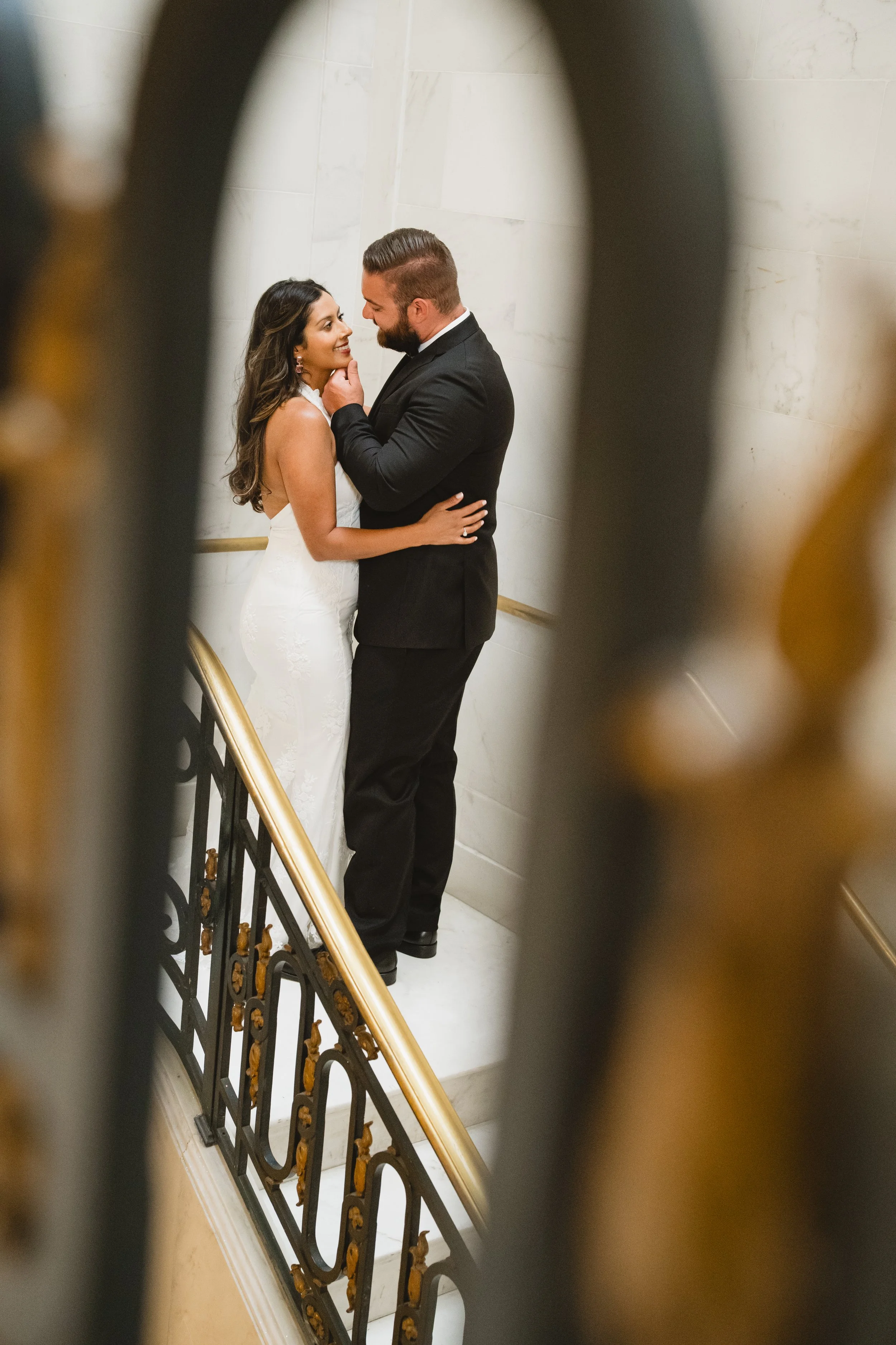 Couple embracing on a staircase, framed through the scrollwork of an ornate iron railing.