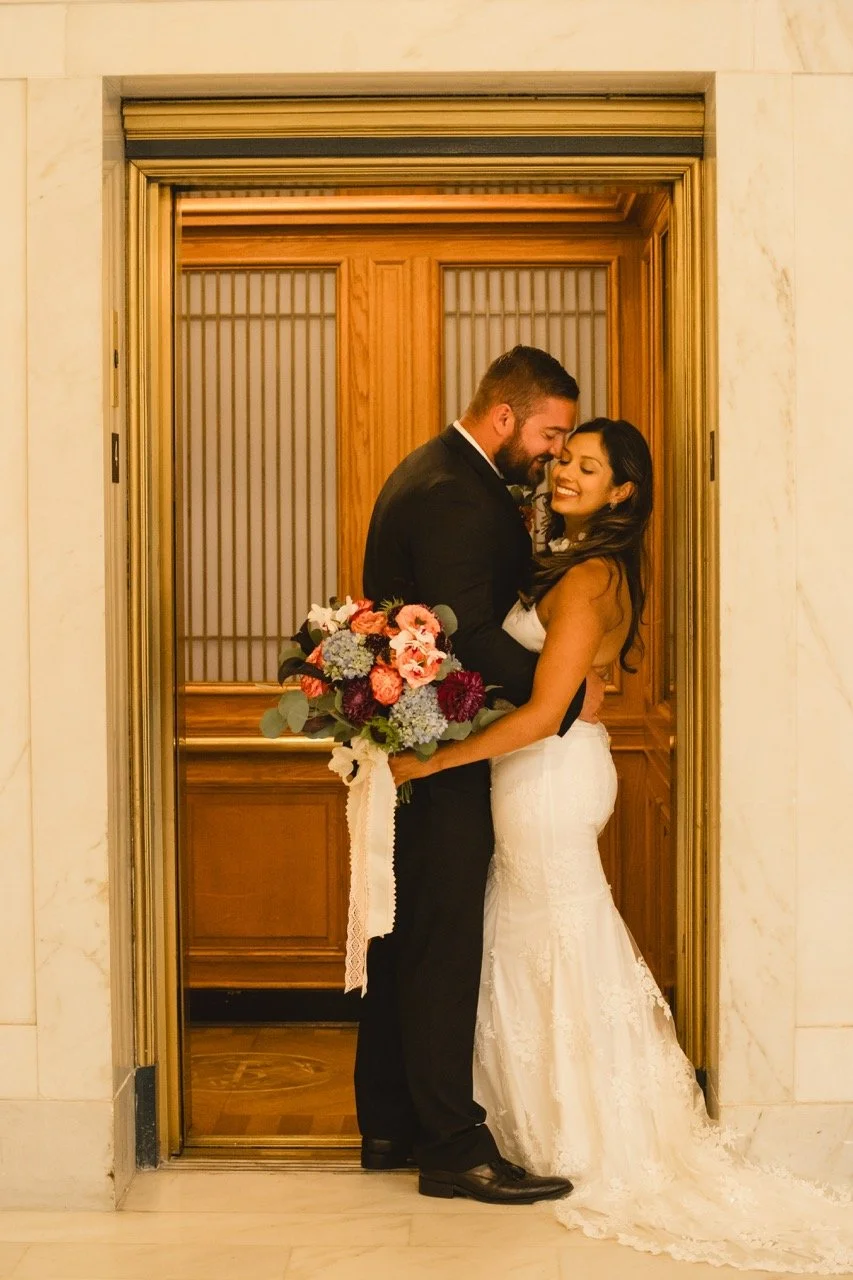 Couple embracing in front of vintage wood-paneled elevator doors, bouquet held in the foreground.