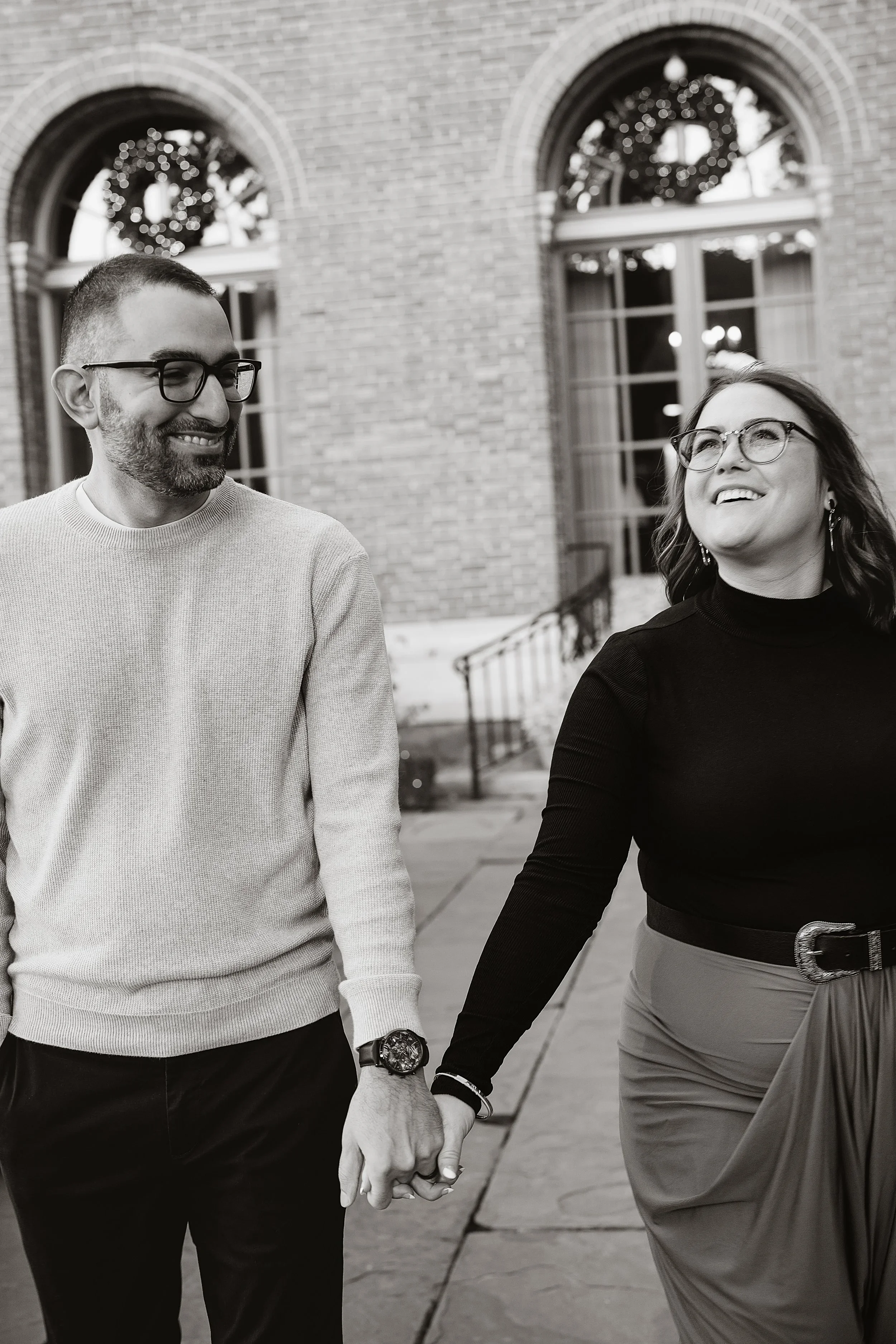Black and white frame of a couple walking toward the camera hand in hand, arms stretched between them, both looking away and laughing.