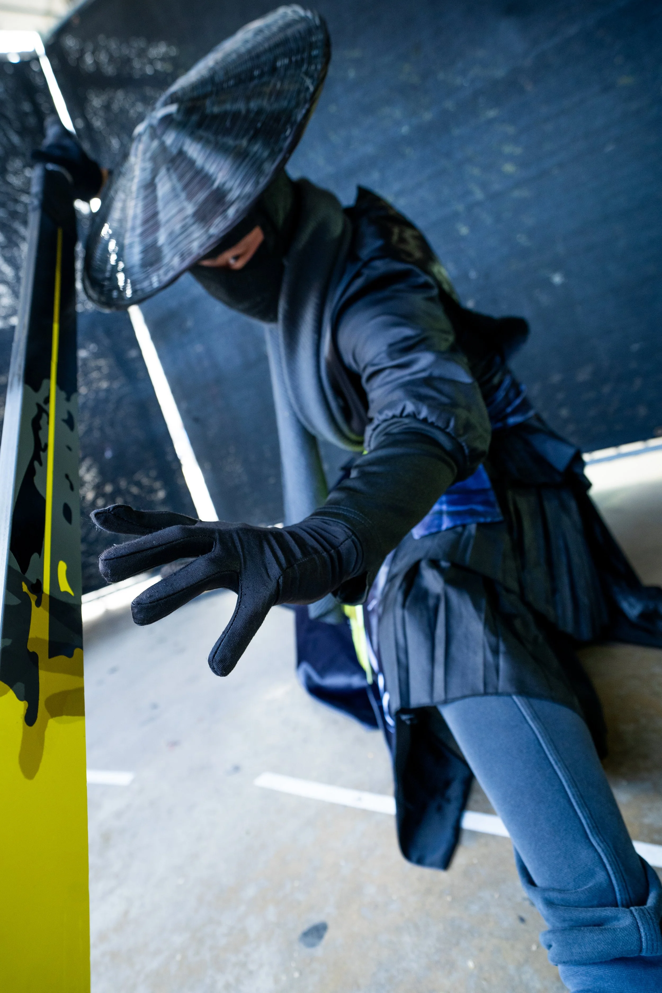 A person wearing a wide-brimmed hat, black face mask, black gloves, and a black jacket is kneeling on the ground, holding onto a yellow pole with black and white graphics, in an outdoor setting.