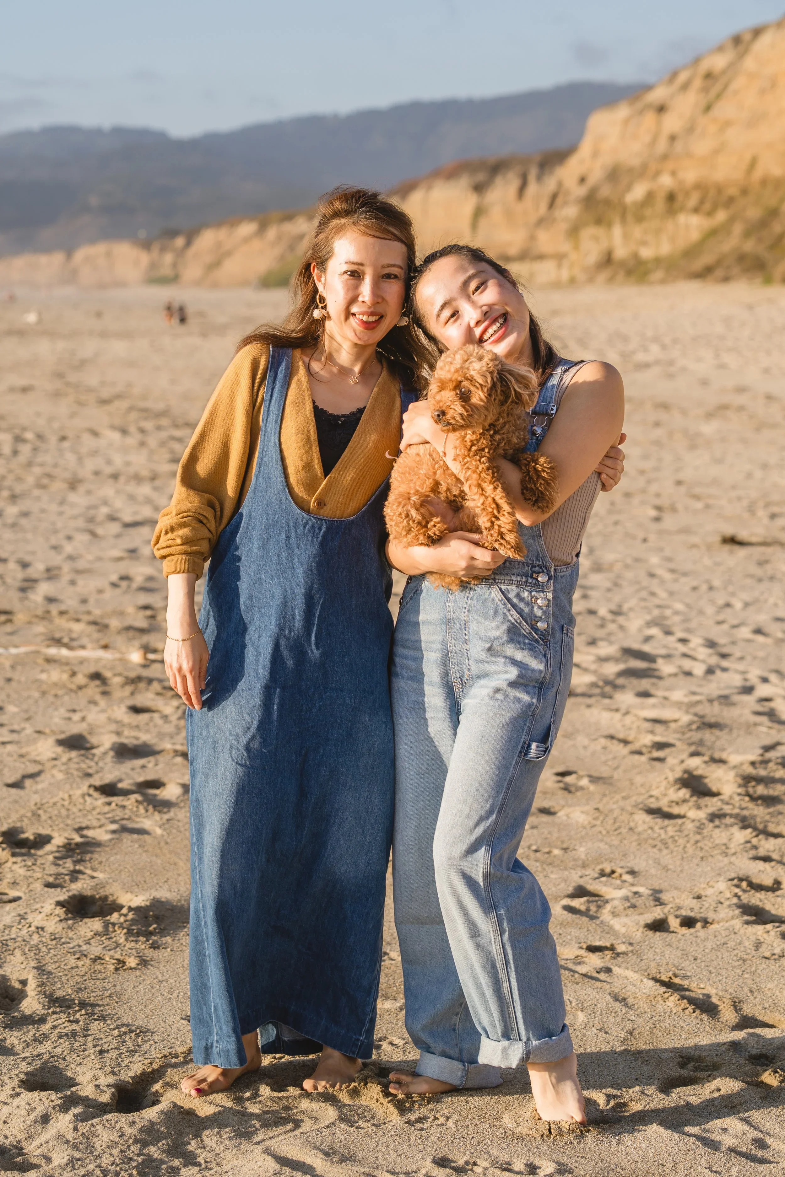 Mother and daughter stand barefoot in denim on the sand, the daughter holding their small poodle mix, cliffs and hills in the distance