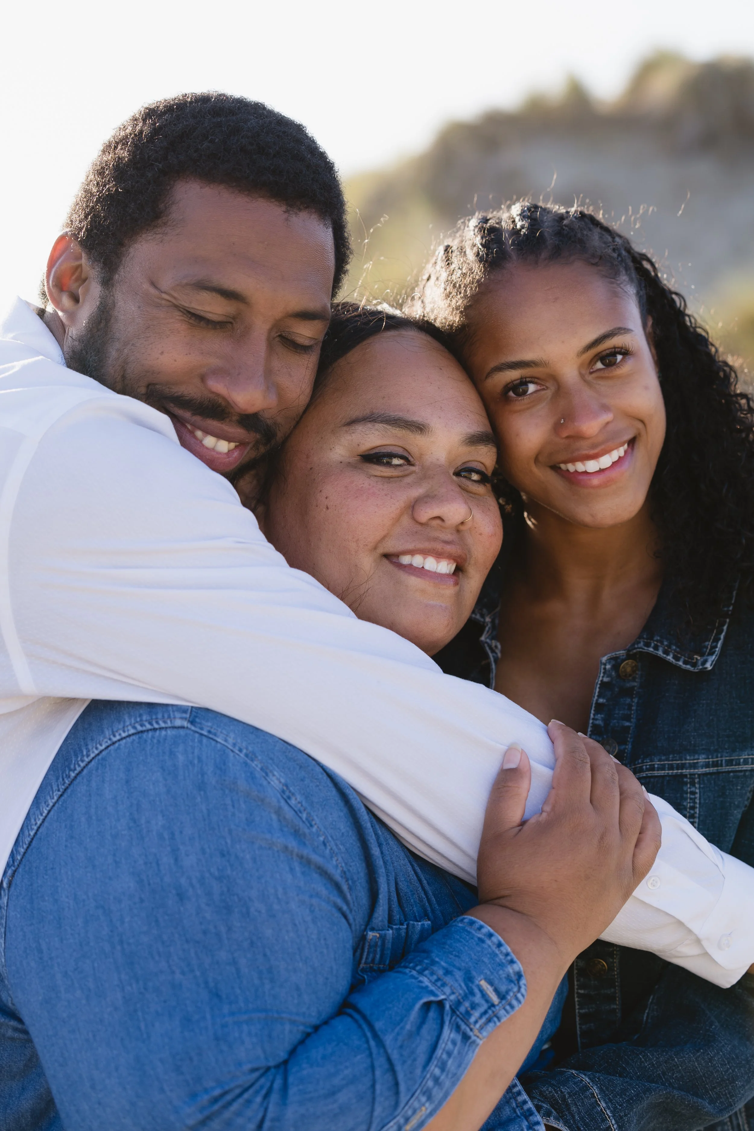 Three friends hugging outdoors, smiling and enjoying a sunny day.