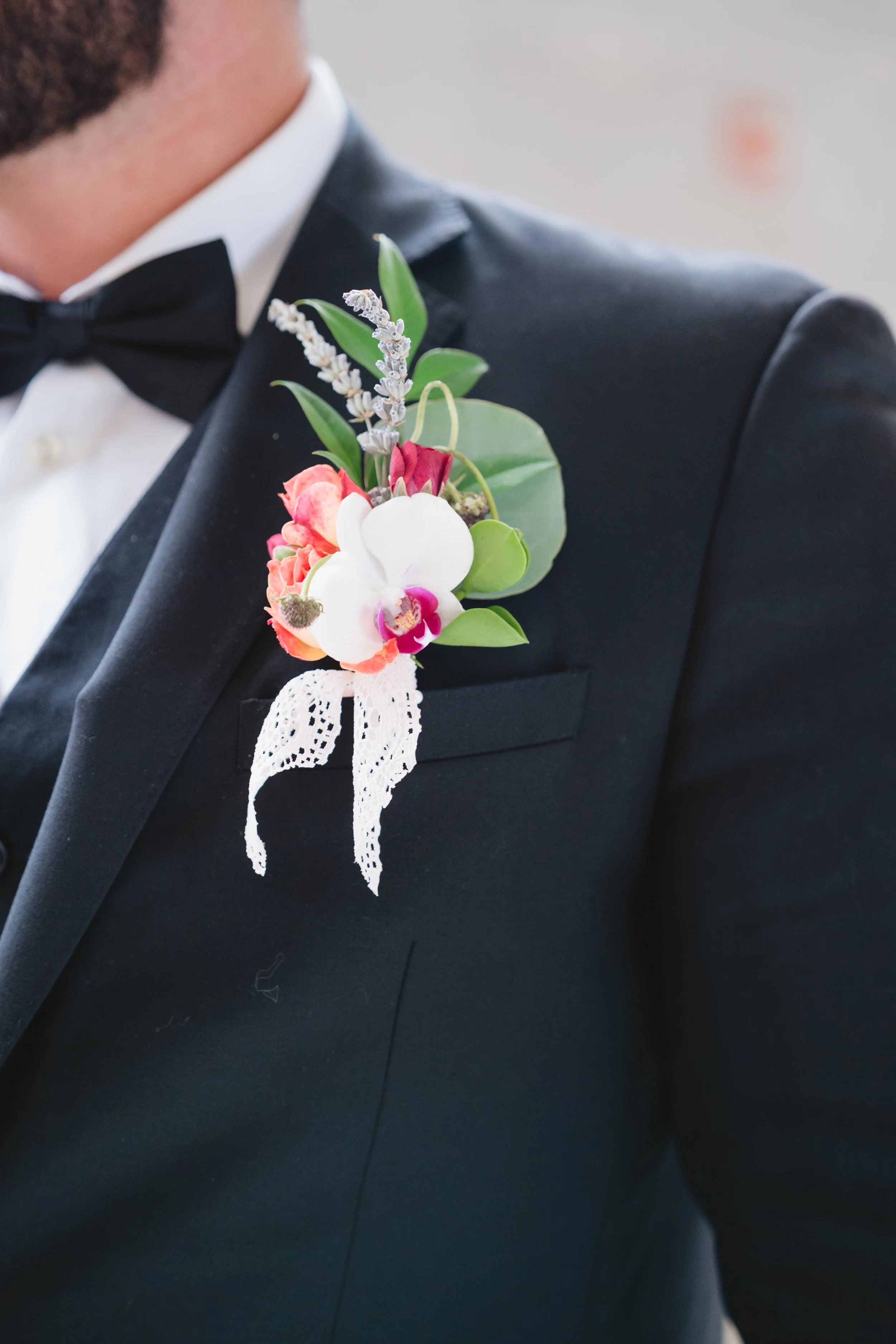 Detail of a boutonniere with orchids, roses, and trailing lace ribbon pinned to a groom's lapel.