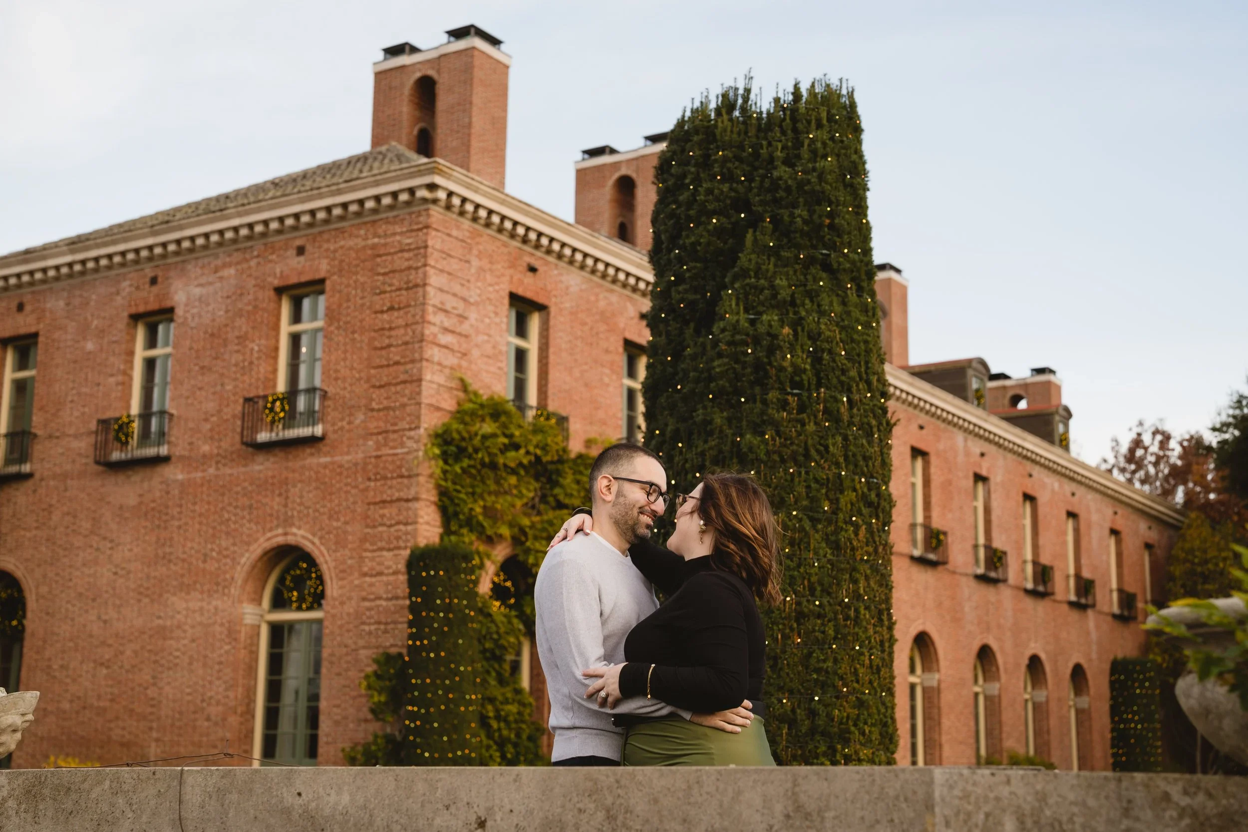 Engagement portrait framed against Filoli's red brick house, with a tall cypress wrapped in lights and wreaths hanging in the windows.