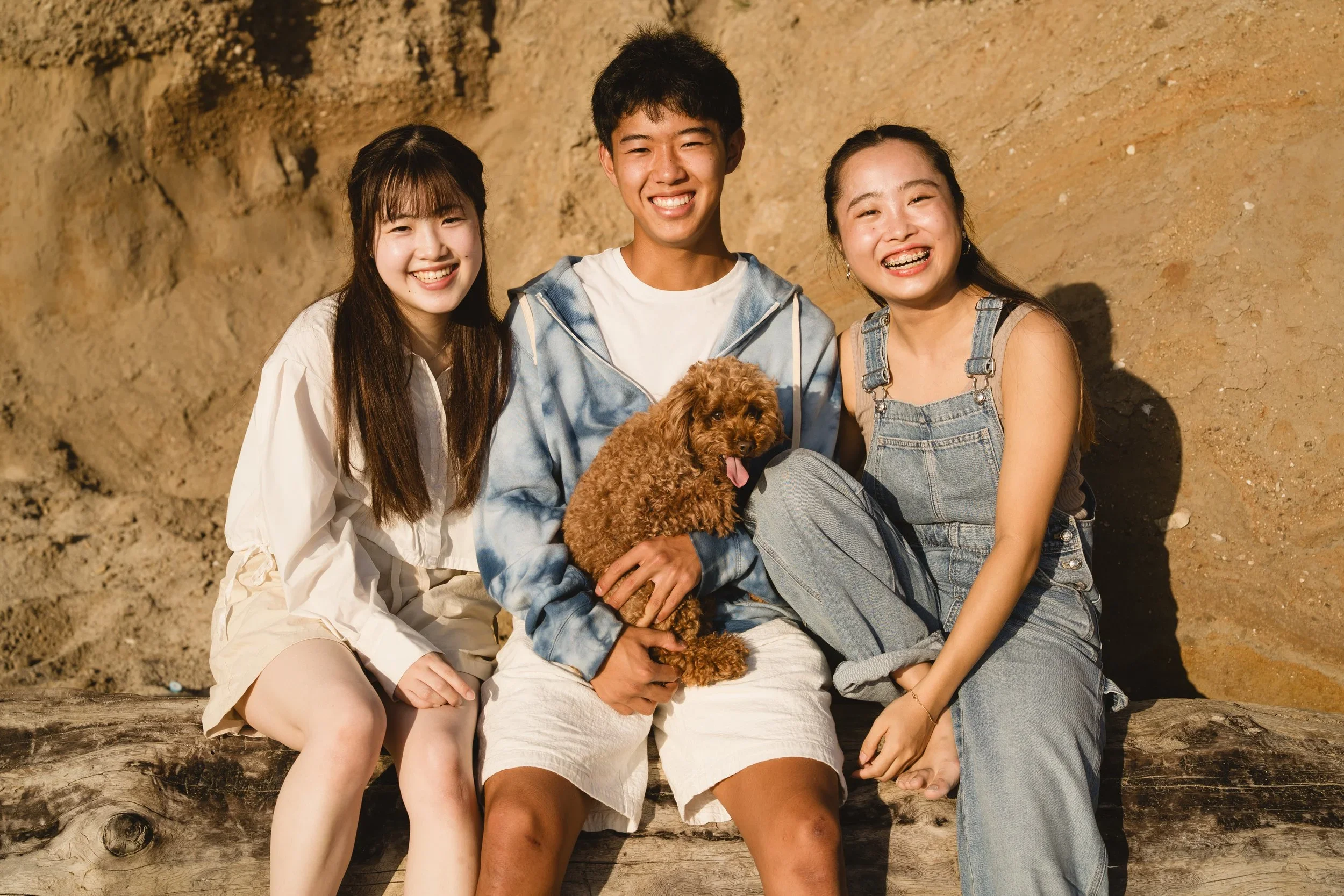 Three siblings sit shoulder to shoulder on a driftwood log at the base of a sandstone bluff, the brother holding their poodle mix on his lap