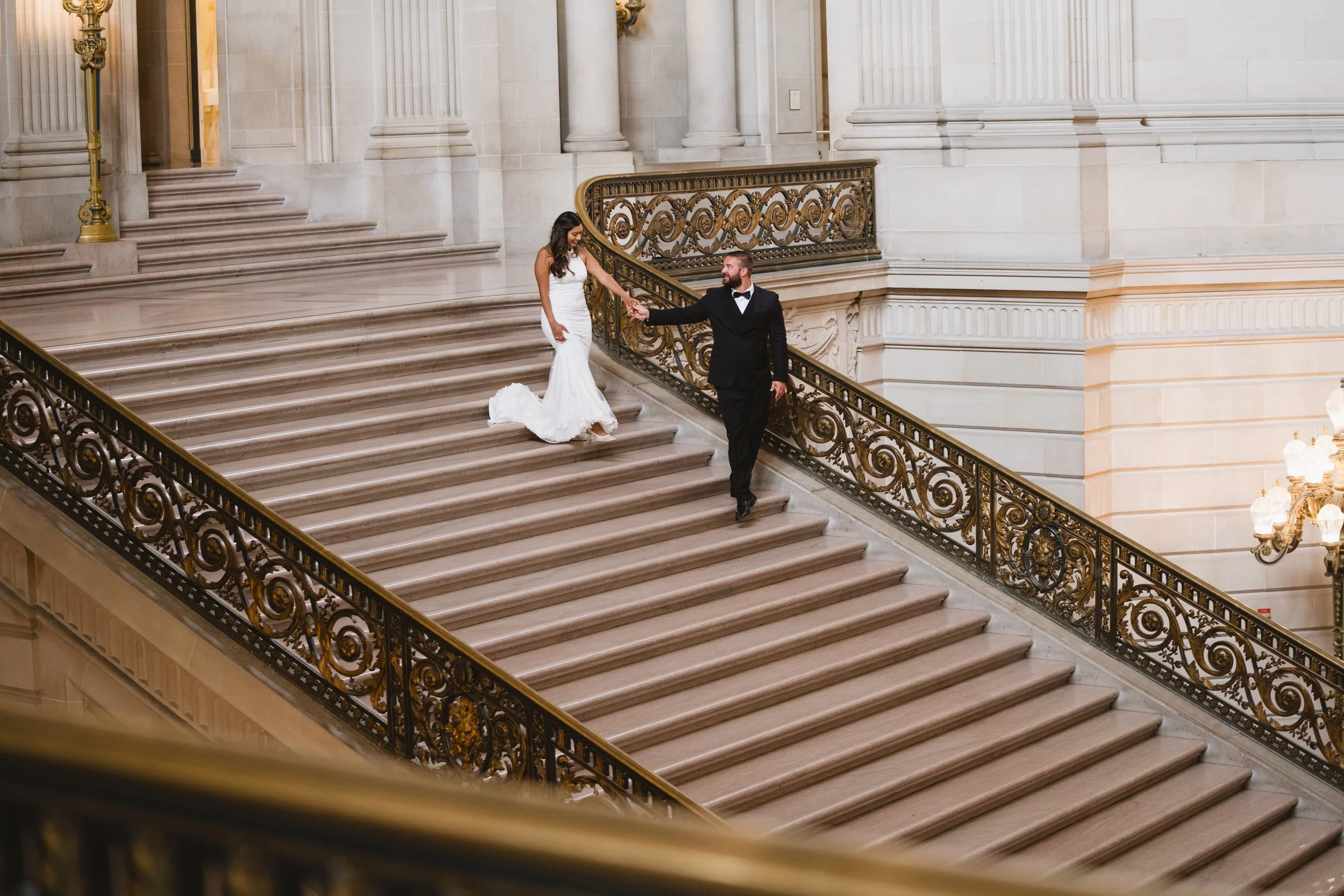 Aerial view of a couple walking hand in hand down an ornate side staircase with gilded railings.