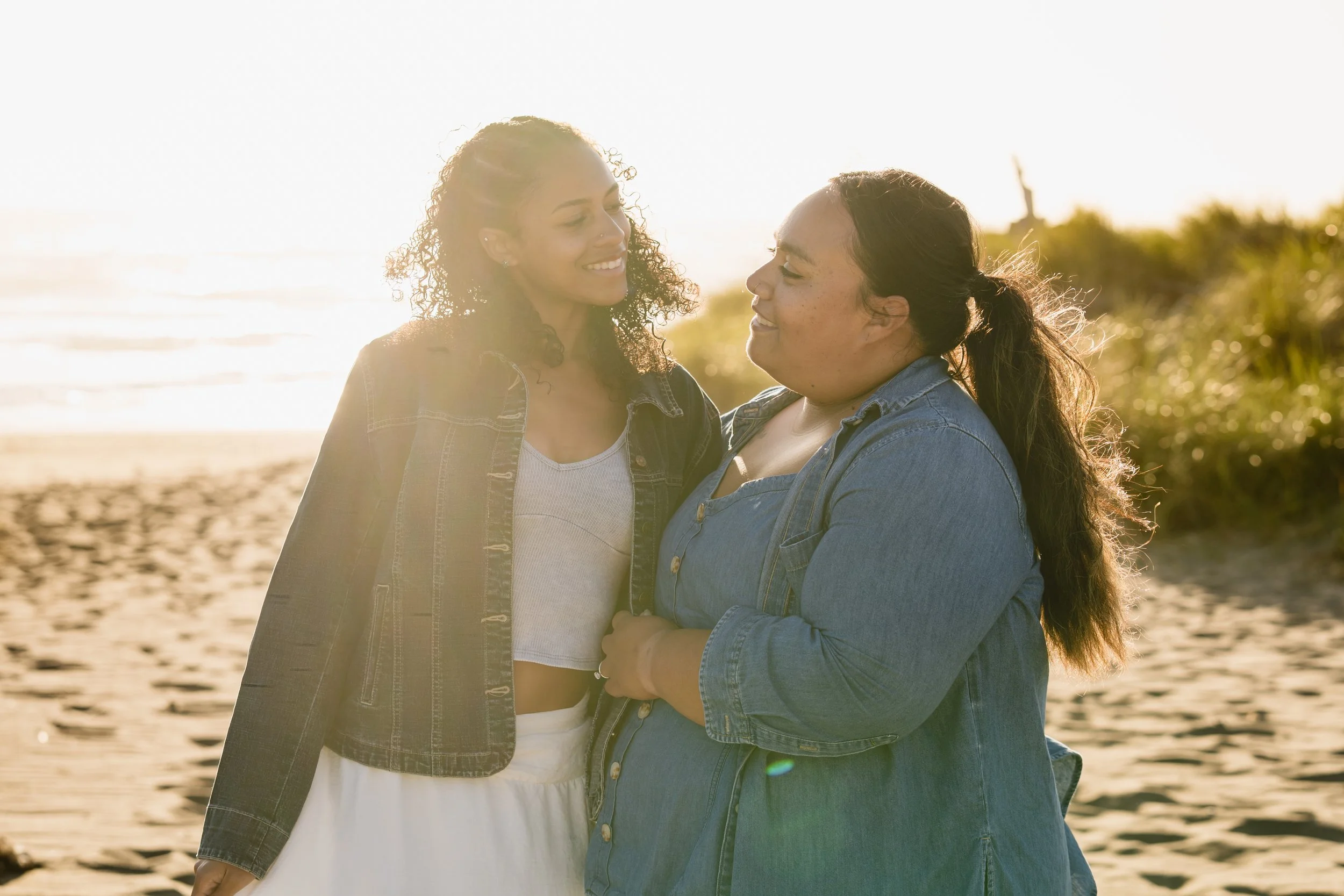 Two women standing close together on a sandy beach in front of the ocean with the Statue of Liberty in the background, during sunset.