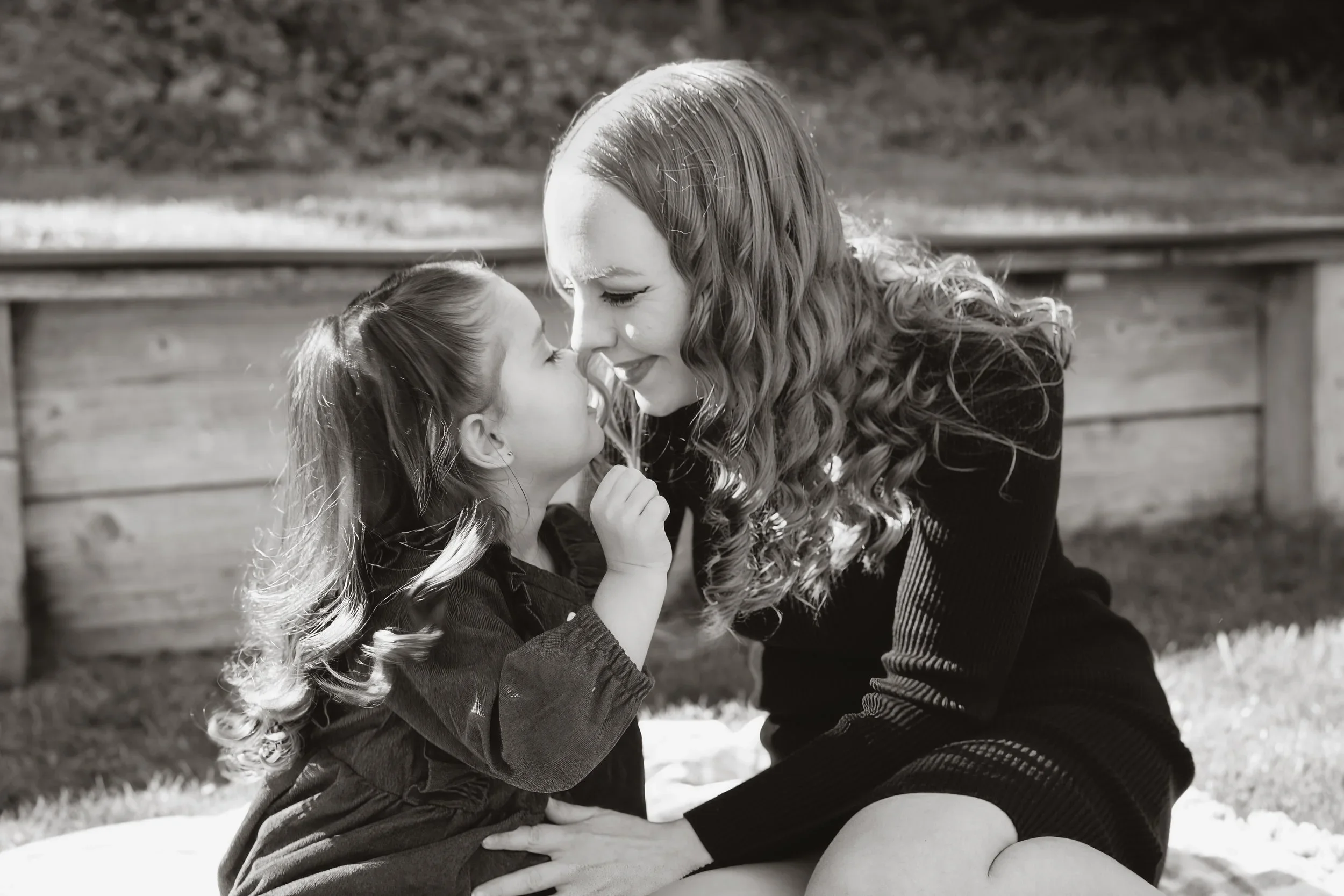 Black and white frame of a daughter leaning in to kiss her mom on the nose while the two sit close together on a blanket outdoors